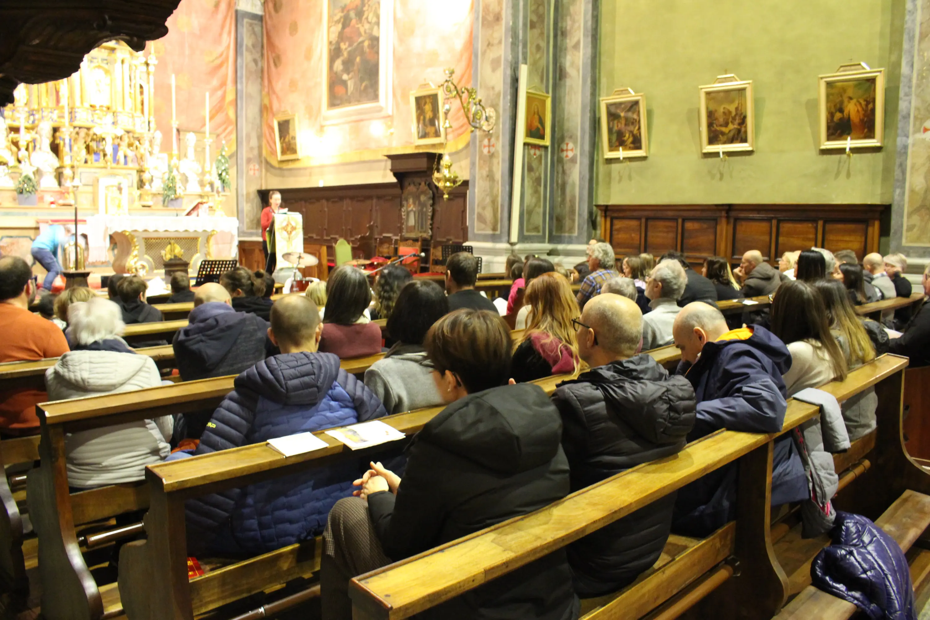 Una vista di una chiesa con molte persone sedute sui banchi. Si possono vedere opere d'arte alle pareti e un altare decorato sullo sfondo. Alcuni partecipanti