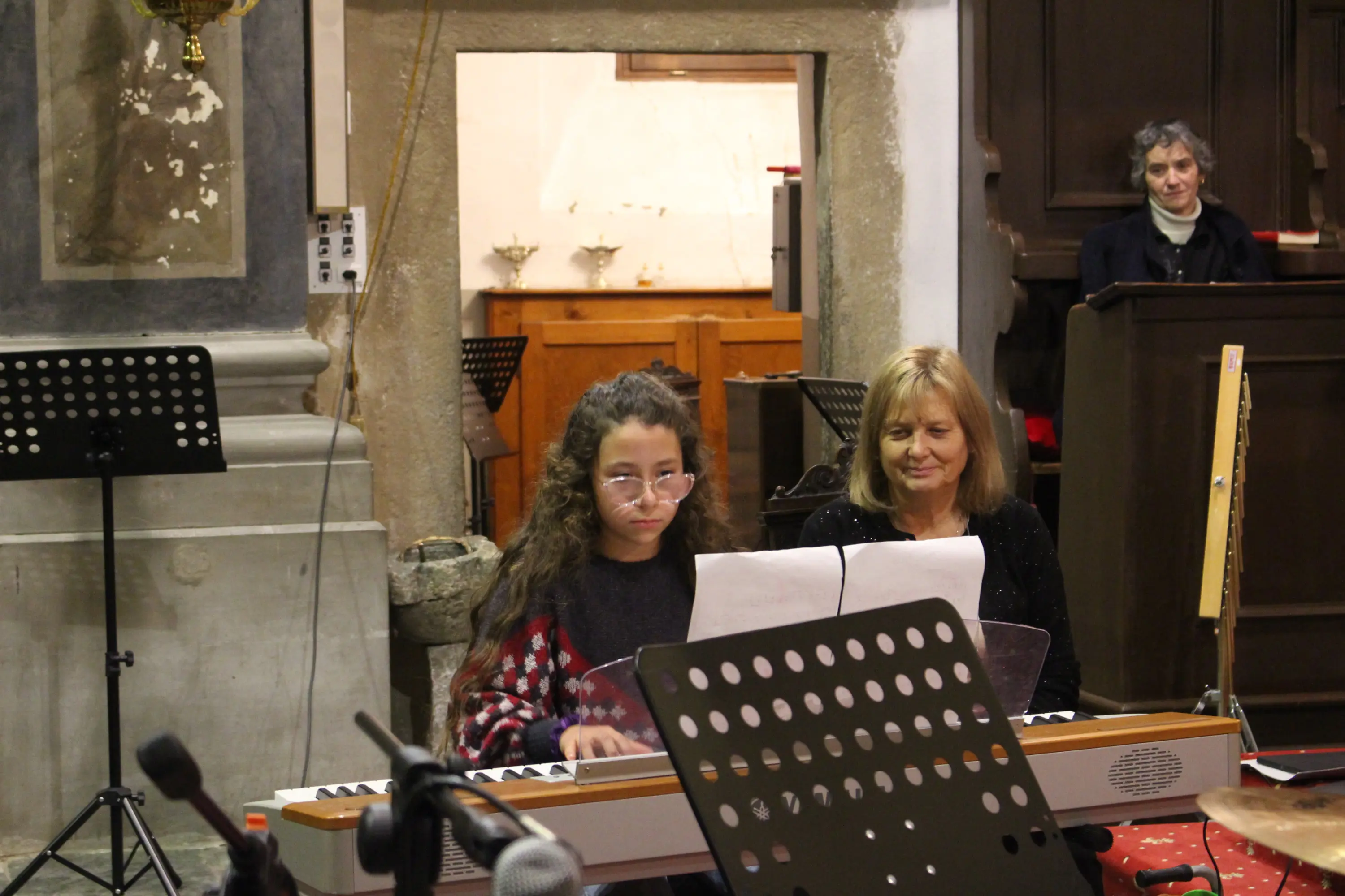 Una ragazza suona il pianoforte mentre una donna la osserva con un sorriso. Sullo sfondo, si intravede un'altra persona che assiste all'esibizione in un