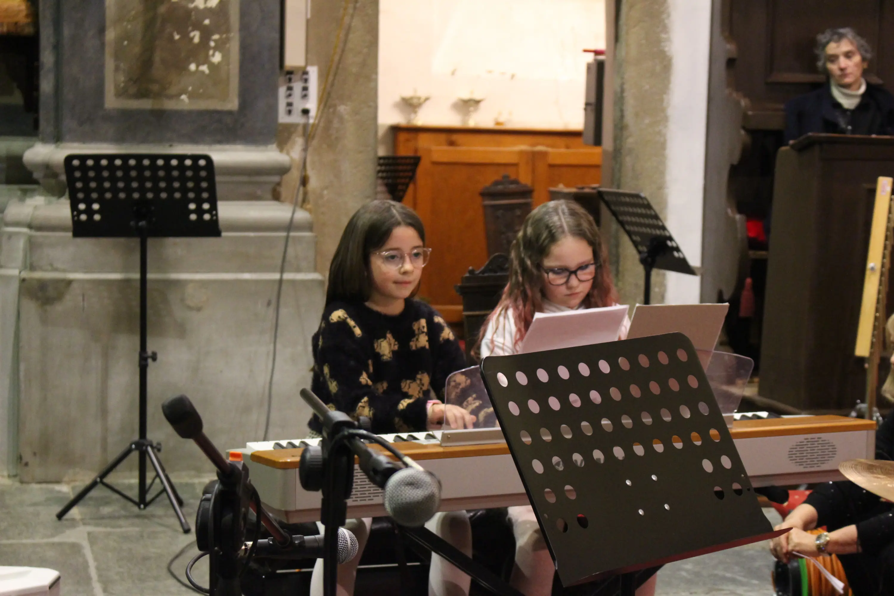 Due ragazze suonano un pianoforte in un contesto di performance musicale. Una delle ragazze indossa occhiali e ha lunghi capelli castani, mentre l'altra ha capelli