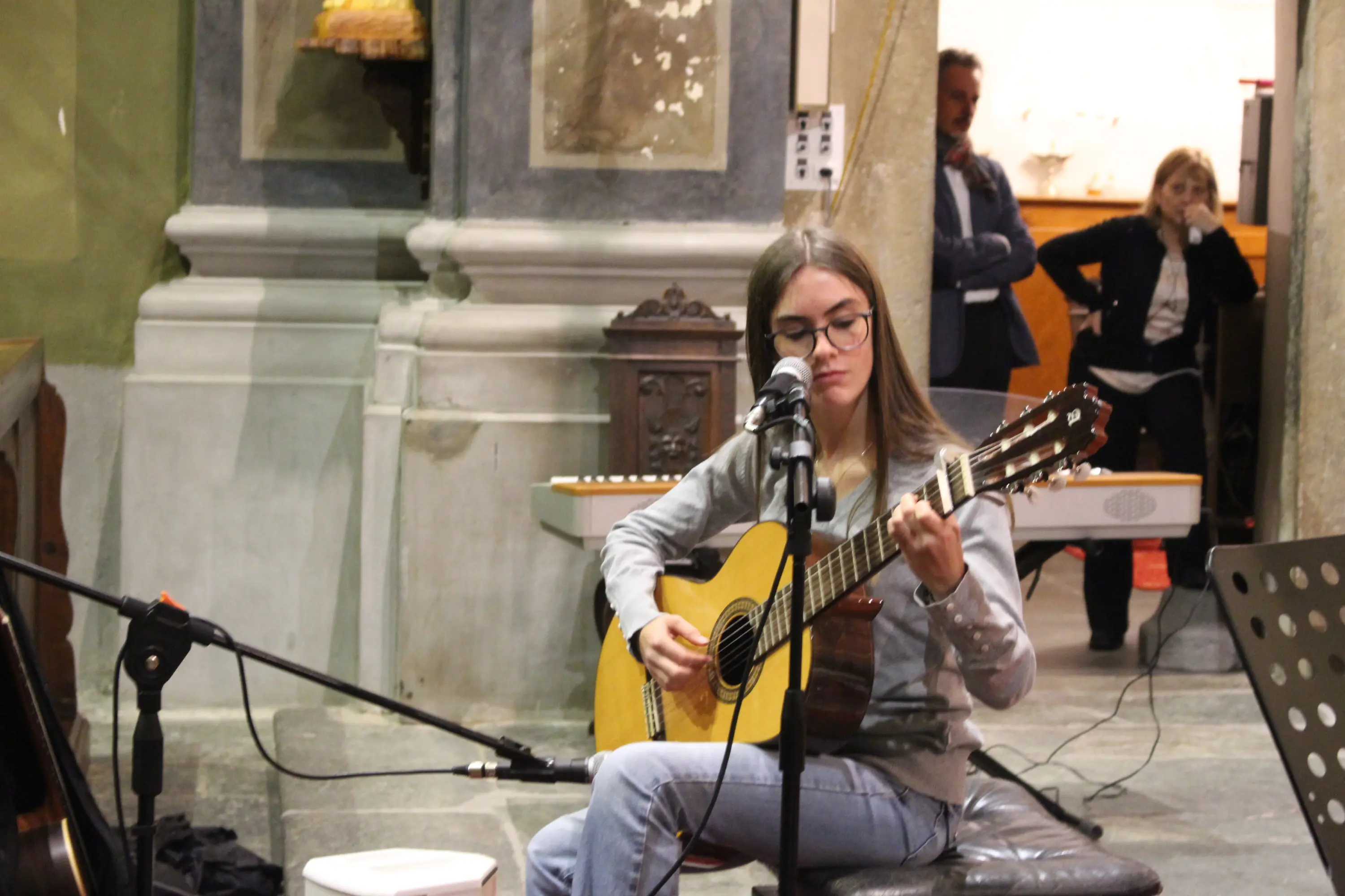 Una giovane donna con occhiali suona una chitarra in un ambiente naturale. È seduta su uno sgabello, concentrata sulla musica. Sullo sfondo si intravede