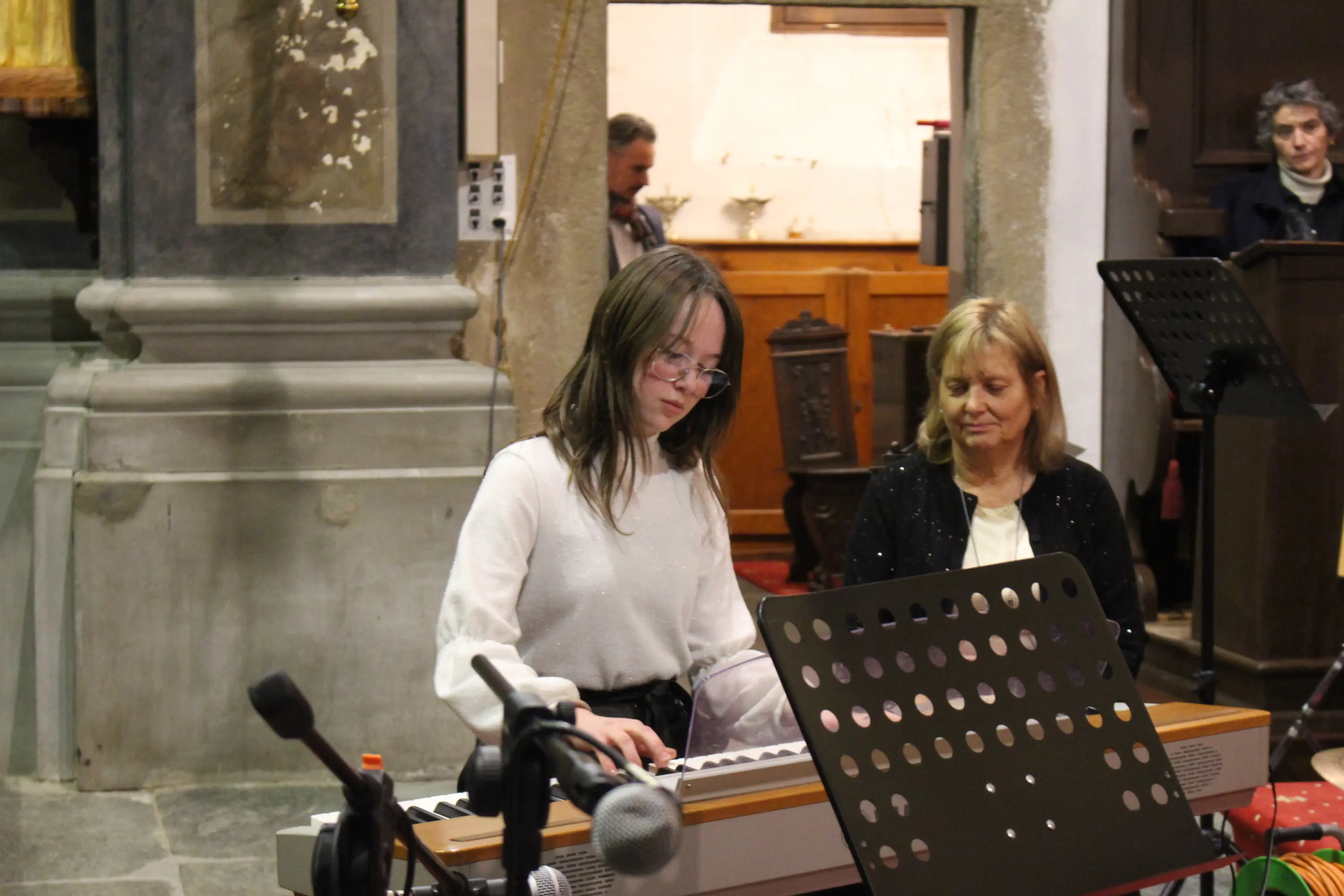 Una giovane musicista suona un pianoforte in una chiesa, mentre un'altra donna la osserva. L'ambiente è accogliente, con dettagli architettonici visibili sul