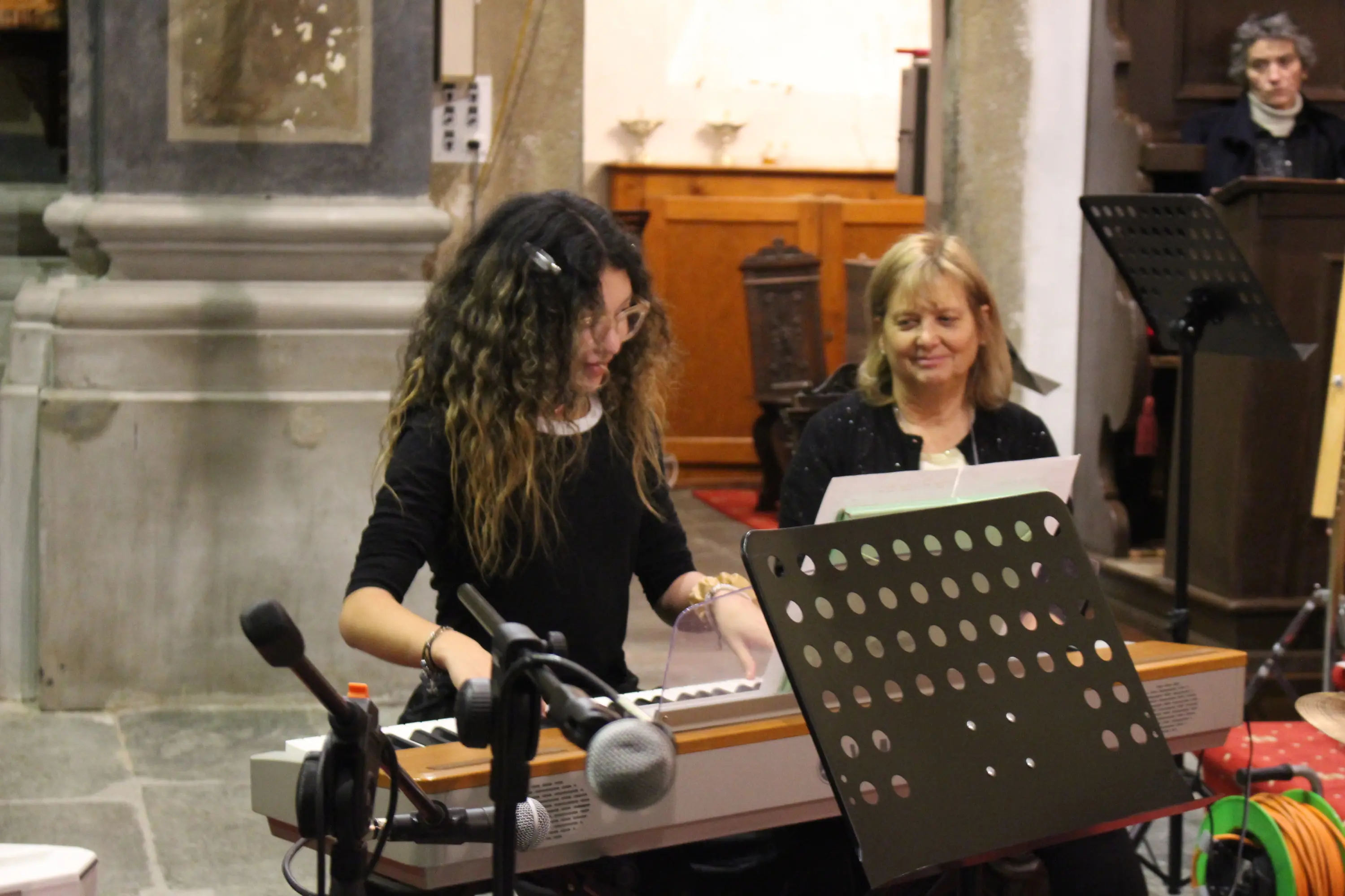 Una giovane musicista suona un pianoforte elettronico, mentre una donna adulta seduta accanto osserva con interesse. Sullo sfondo, un ambiente di chiesa con elementi di ar
