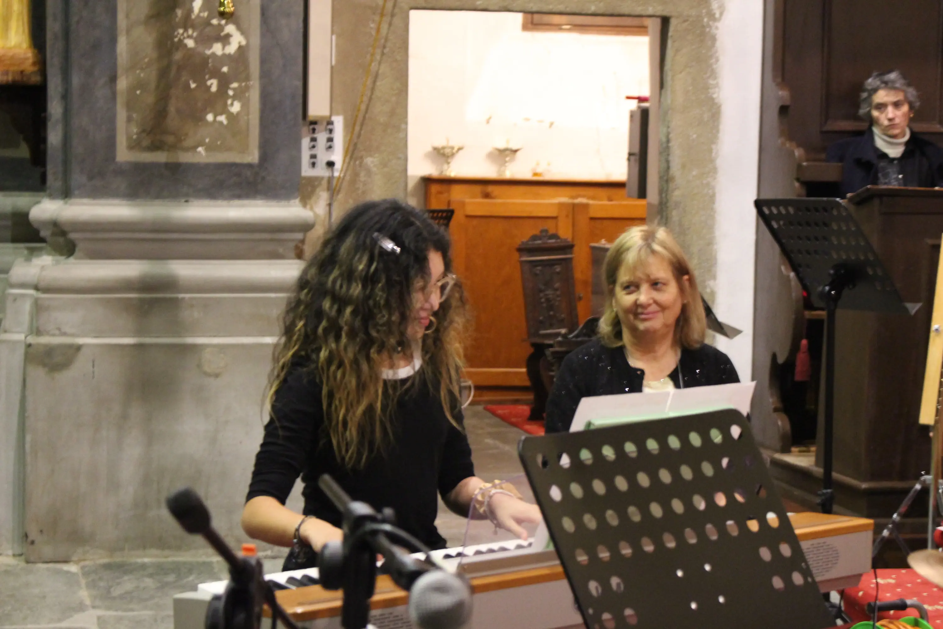 Due donne in una chiesa. Una giovane ragazza suona un pianoforte, mentre un'altra donna adulta sorride guardandola. Sullo sfondo, si intravede un'altra