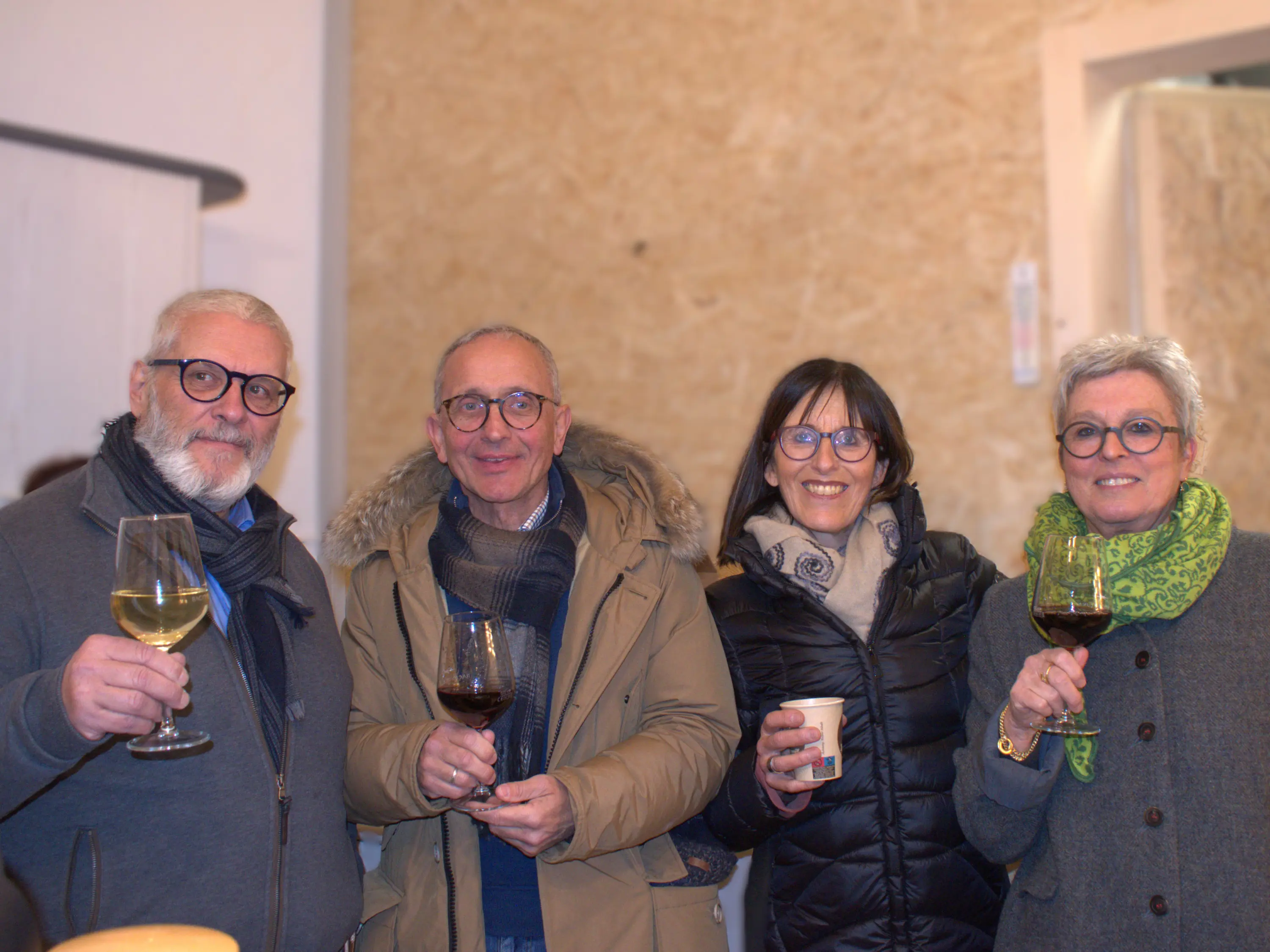 Quattro persone sorridenti tengono bicchieri di vino e un bicchiere di carta in un ambiente accogliente. Sono vestite in modo elegante e informale, con sciarpe