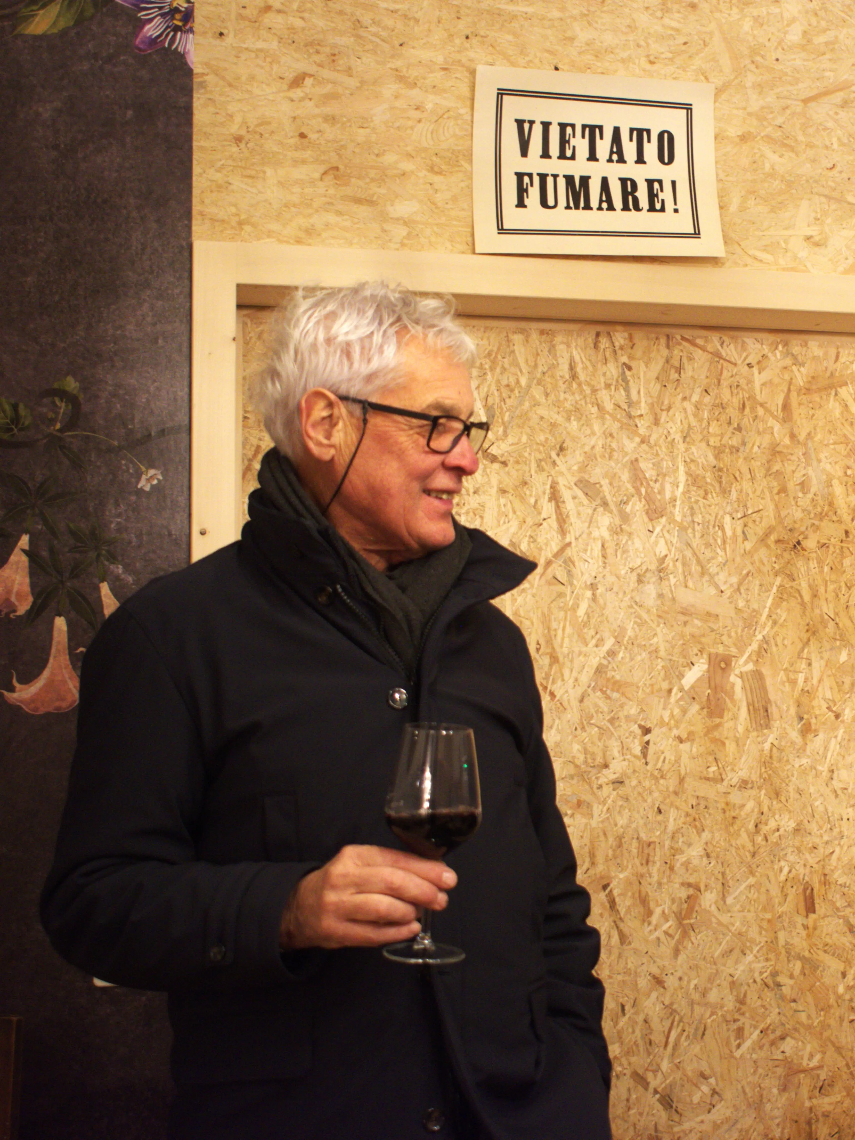 Uomo con capelli grigi e occhiali, in piedi mentre tiene un bicchiere di vino rosso. Sullo sfondo è visibile un cartello che dice "VIETATO