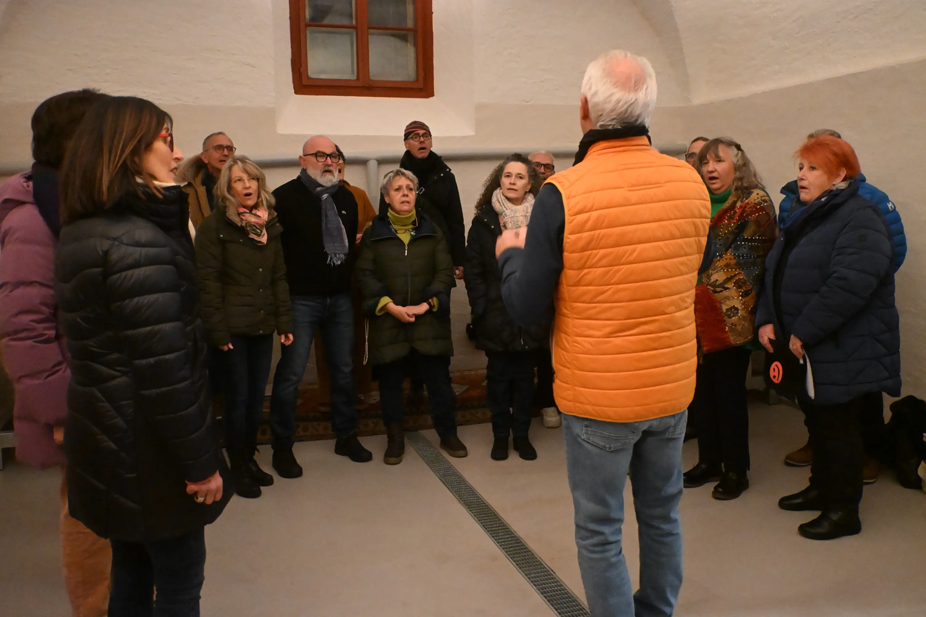 Un gruppo di persone canta in una stanza, accompagnato da un uomo che dirige. Gli adulti indossano giacche e cappotti invernali e sembrano concentrati mentre partecipano