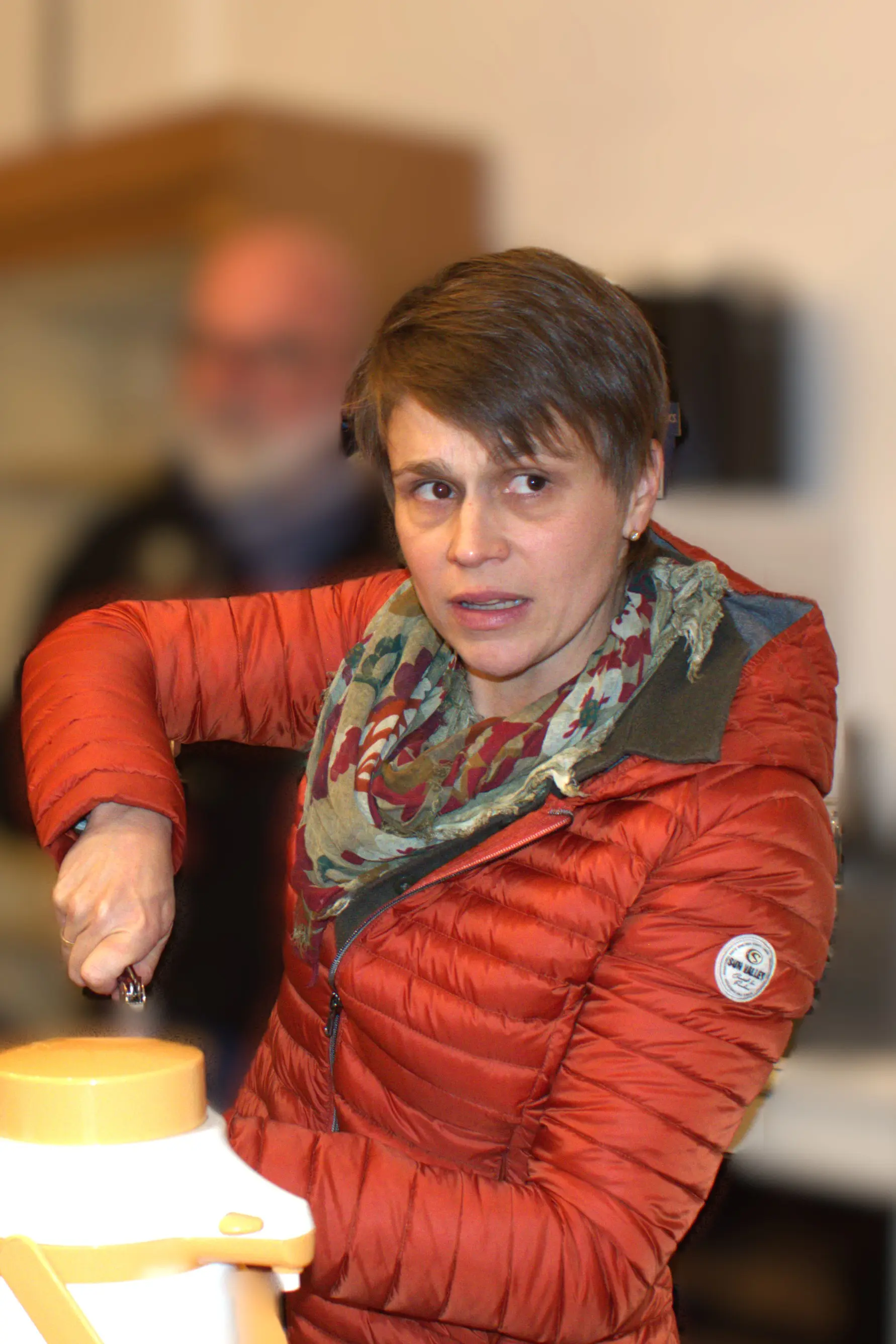 Una donna con giacca rossa e sciarpa a fiori sembra concentrata mentre apre un contenitore, con un'espressione di curiosità o confusione. Sullo sfondo