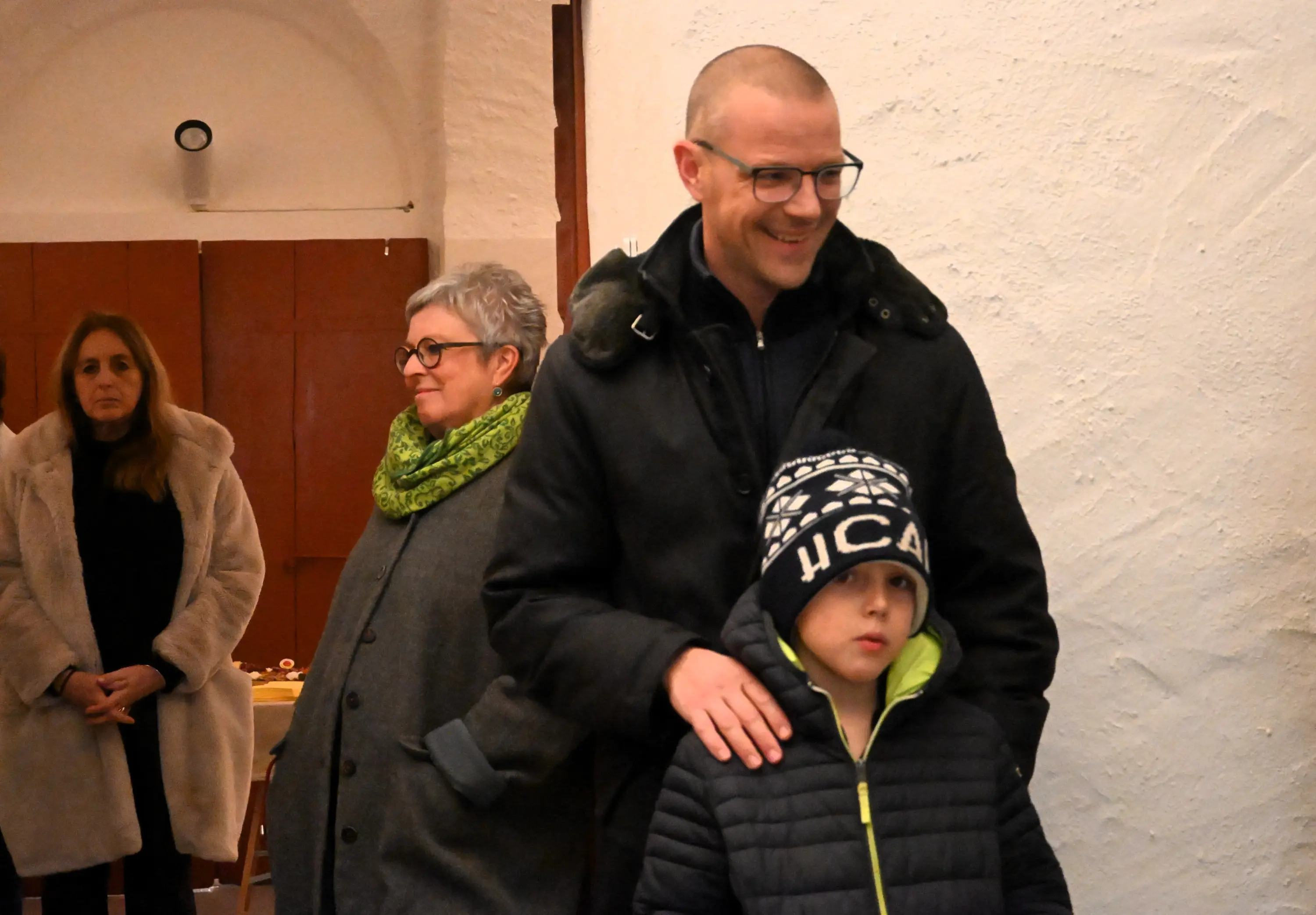 Un uomo sorridente con occhiali e una giacca nera, insieme a un bambino in un giubbotto verde scuro e cappello, si trovano in una stanza con pare