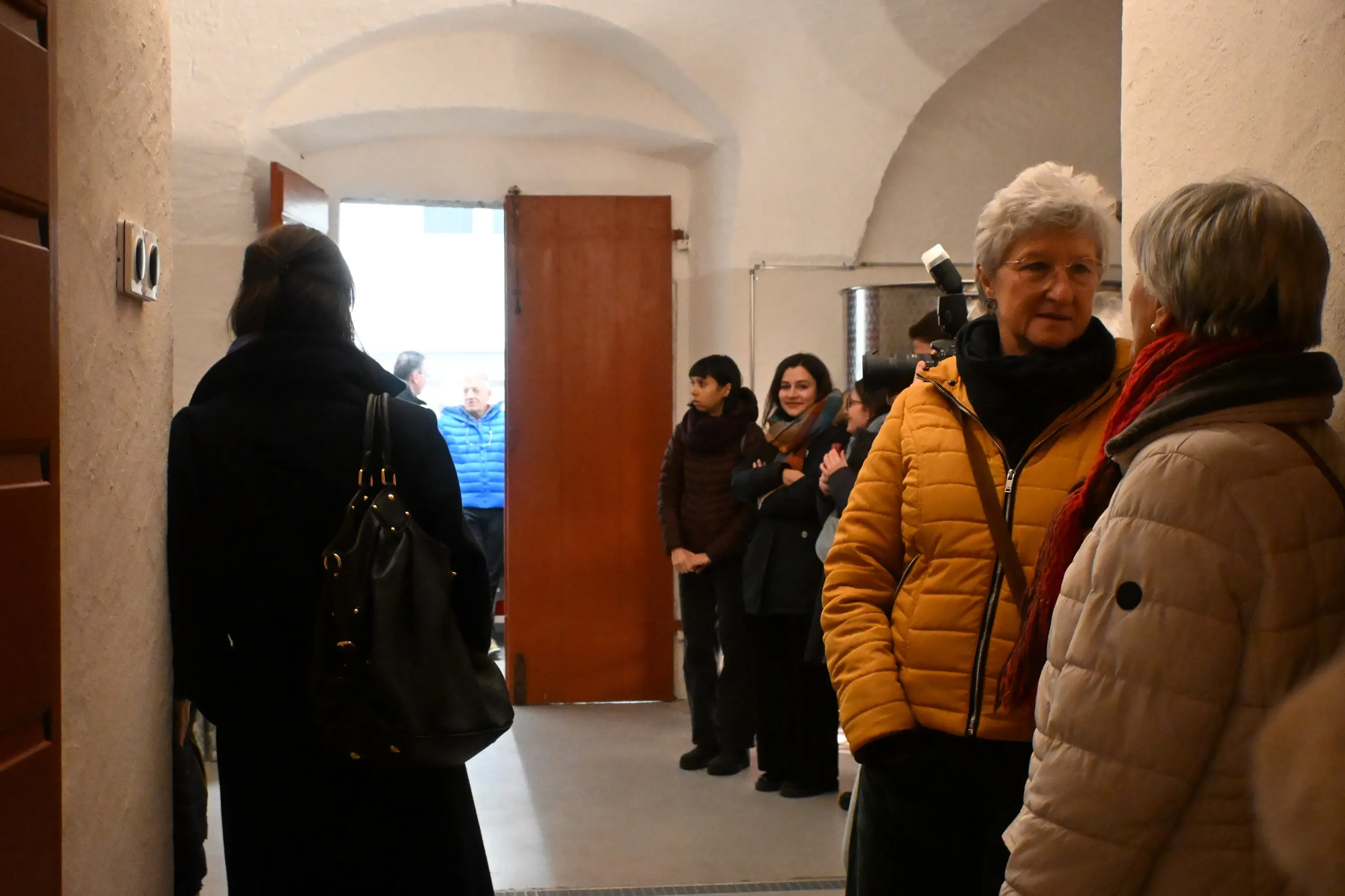 Due donne conversano in un corridoio, mentre altre persone attendono in lontananza. Il corridoio ha pareti bianche e una porta di legno aperta che lascia