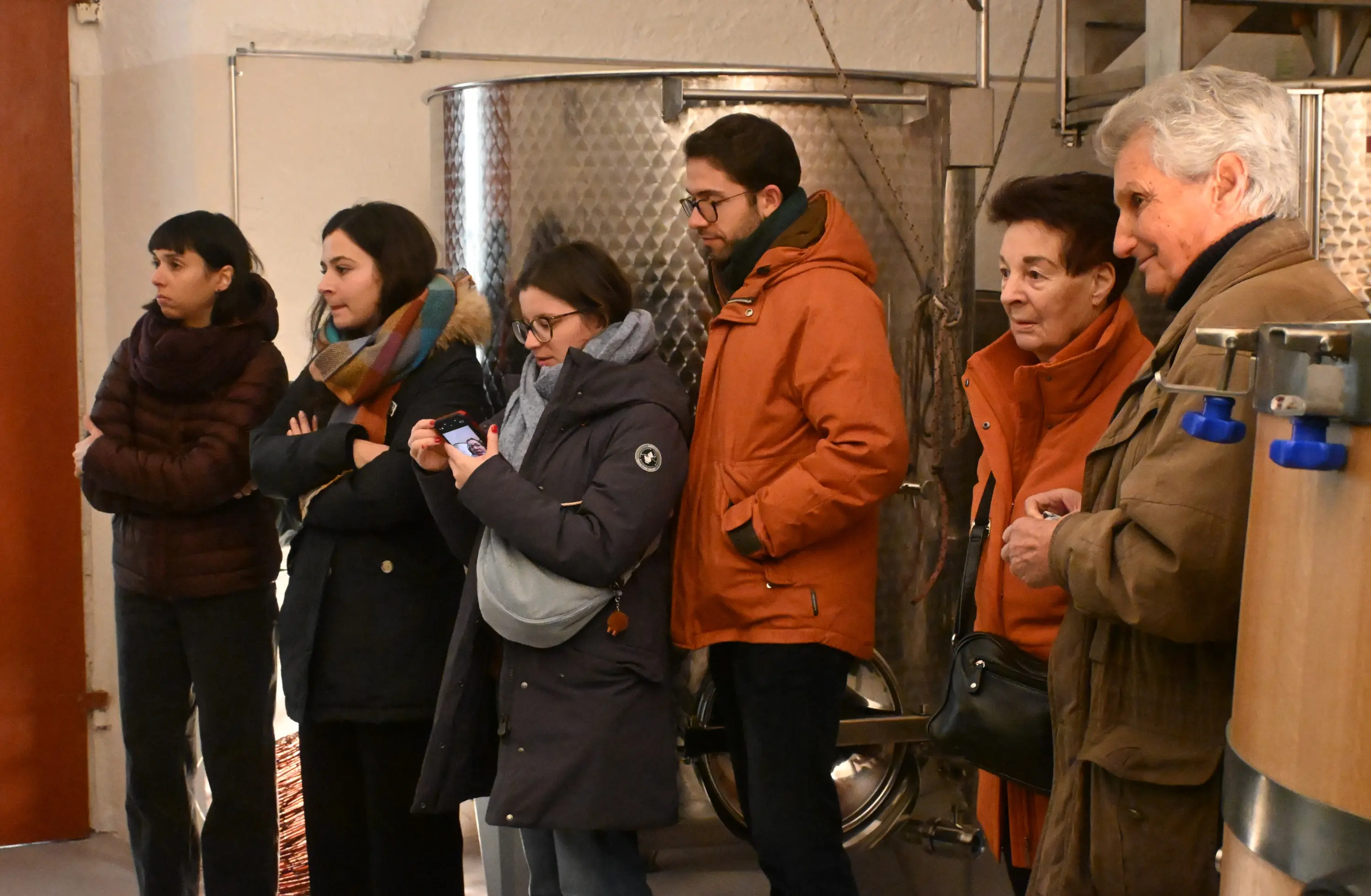 Gruppo di sei persone in una cantina. Alcuni guardano con attenzione, altri controllano i telefoni. Tutti indossano abbigliamento invernale.