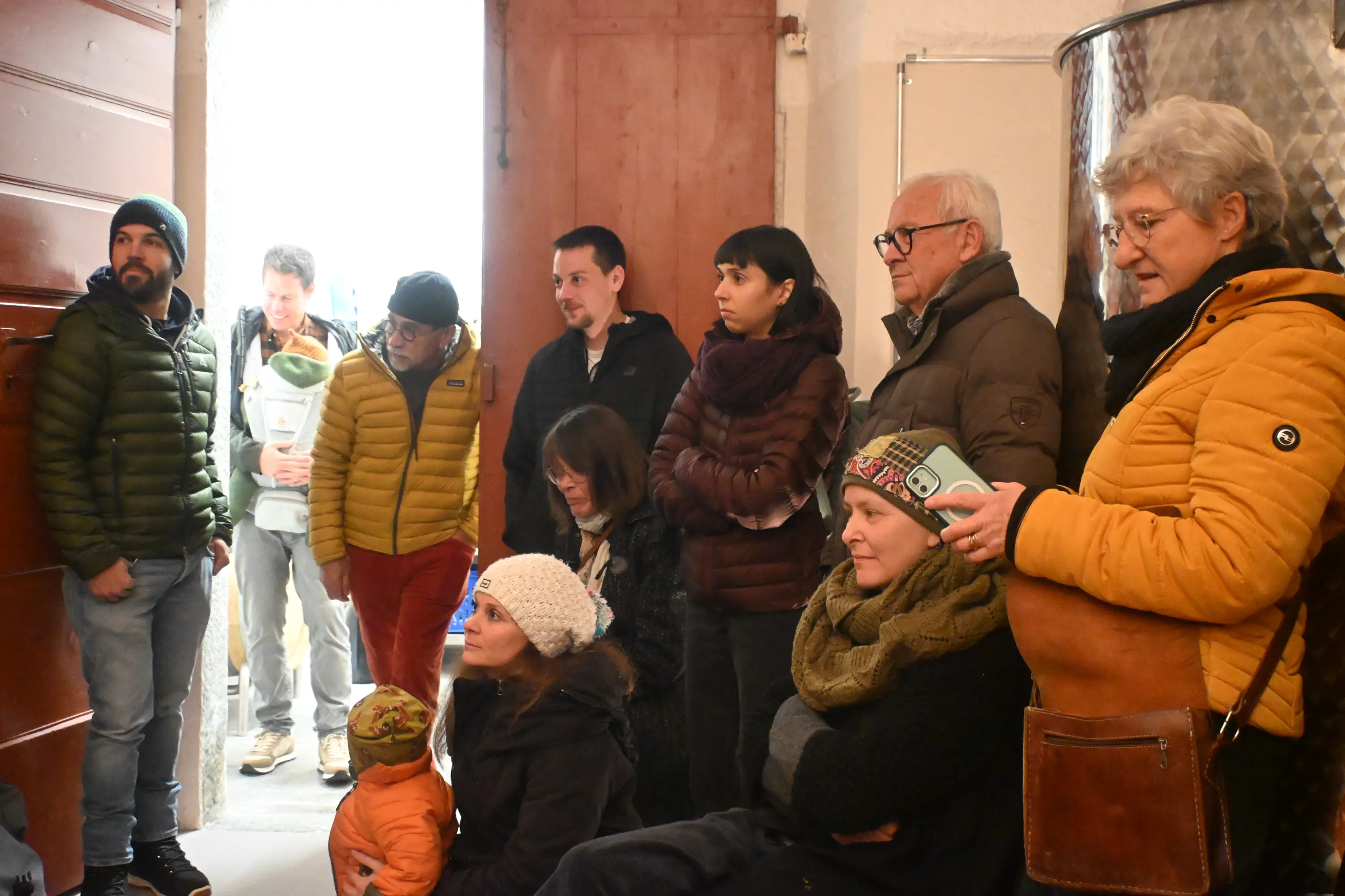 Gruppo di persone in una stanza, alcune sedute e altre in piedi, che ascoltano attentamente. Alcuni indossano giacche pesanti e cappelli, mentre altri hanno