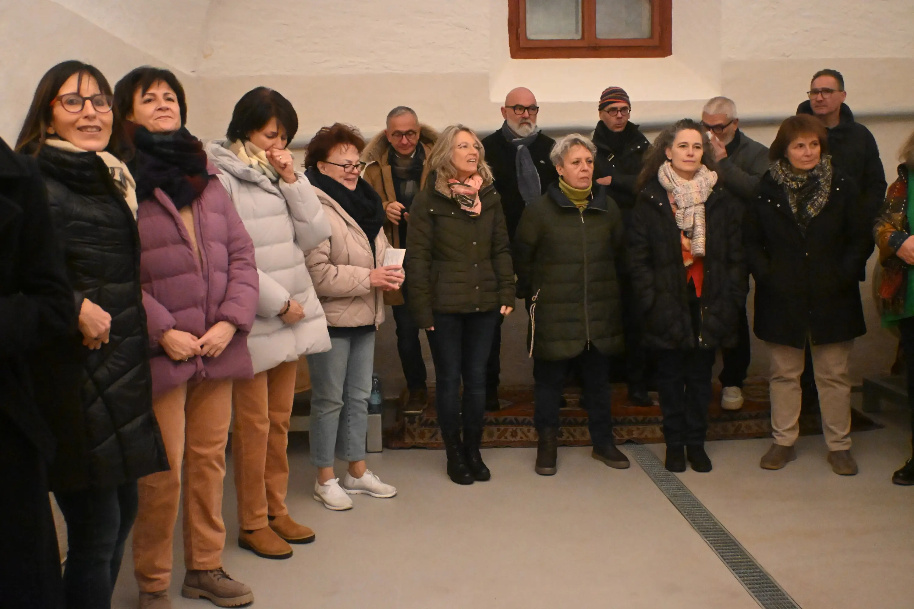Gruppo di persone in una stanza con pareti chiare, sorridenti e in piedi in fila. Alcuni indossano giacche pesanti e sciarpe, mentre altri hanno ab