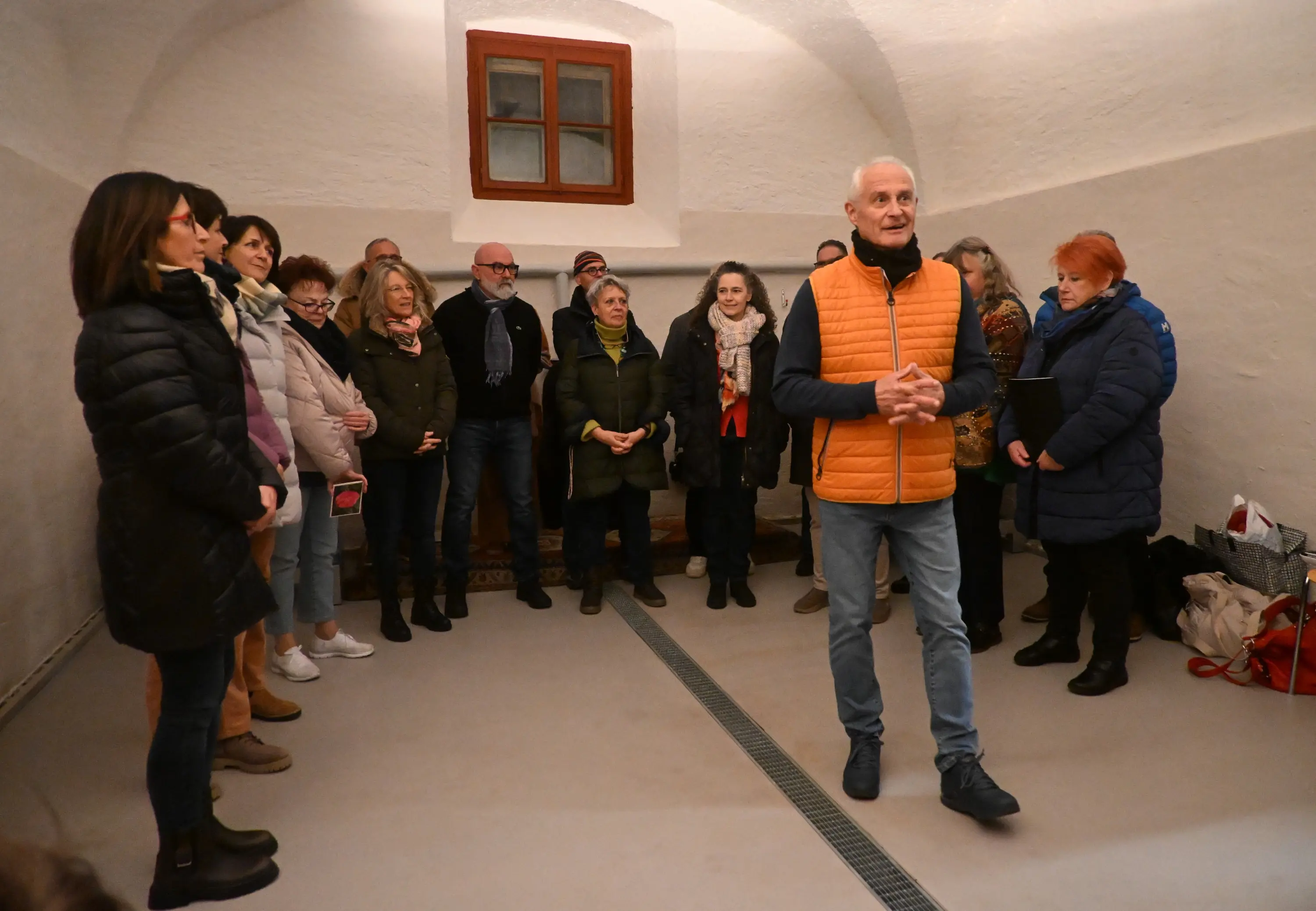Un gruppo di persone si trova in una stanza con pareti bianche e un soffitto a volta. Un uomo in giacca arancione sta parlando, mentre gli altri, in ab