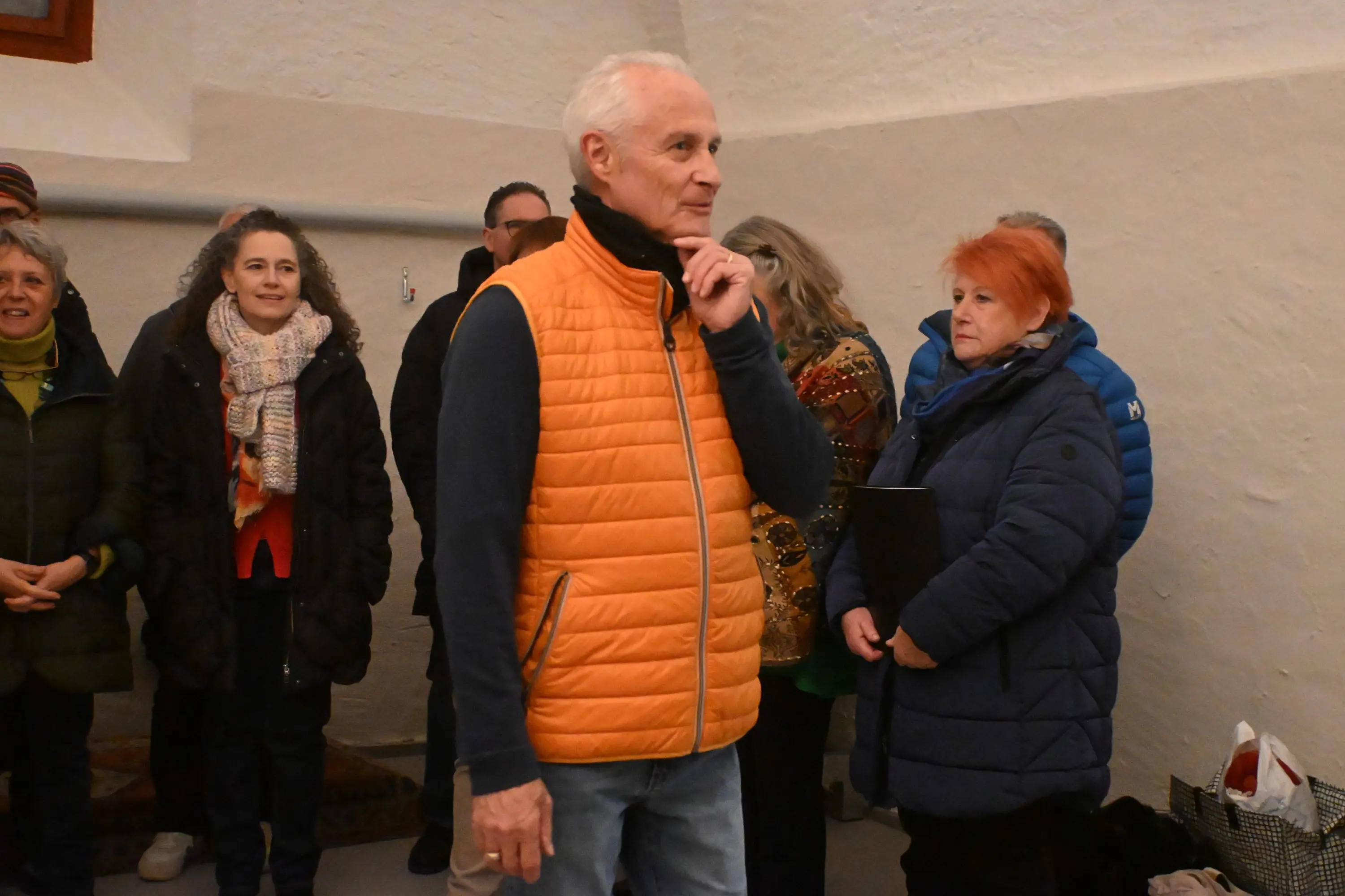 Un uomo con un gilet arancione parla mentre un gruppo di persone lo ascolta in una stanza. Una donna con capelli rossi guarda attentamente e altre persone sono in background, sorridenti