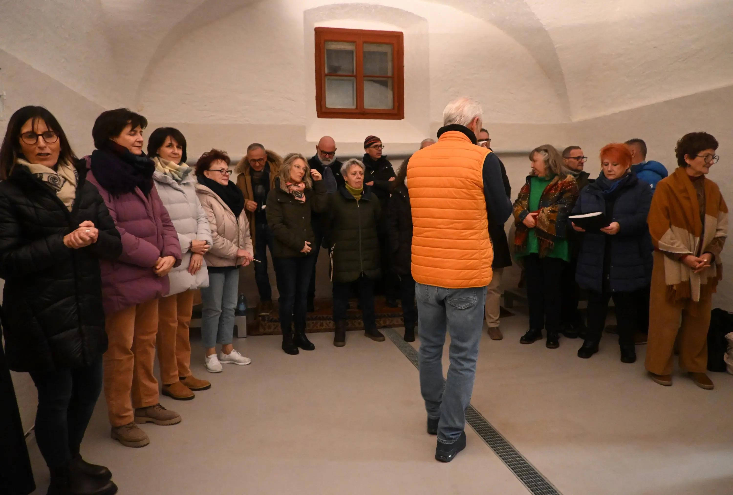 Gruppo di persone in una stanza con pareti bianche, alcune in posa per una foto, altre di spalle. Un uomo con un gilet arancio si volta verso il gruppo mentre