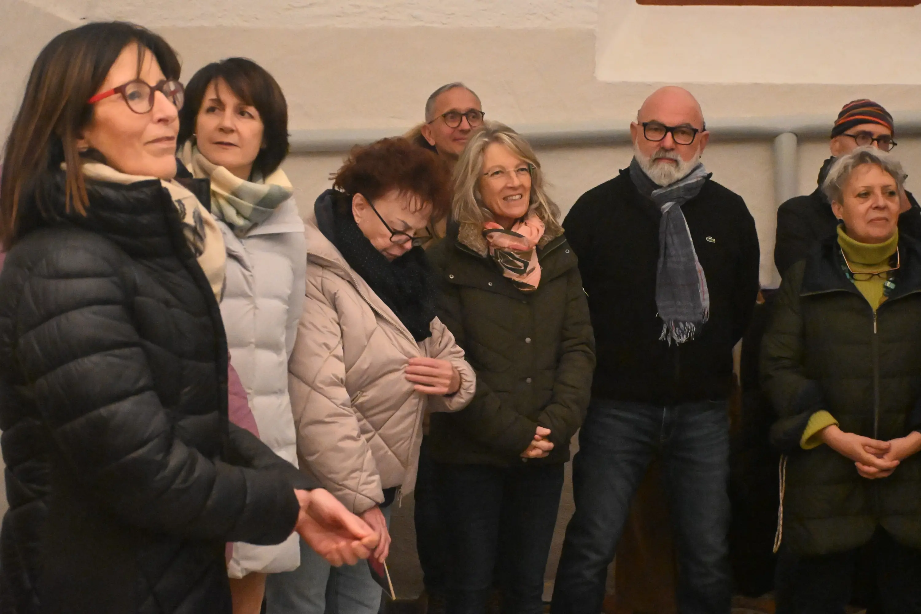 Gruppo di persone in piedi, sorridenti e ascoltando attentamente, in un ambiente interno. Le donne indossano sciarpe e giacche, mentre gli uomini hanno occh