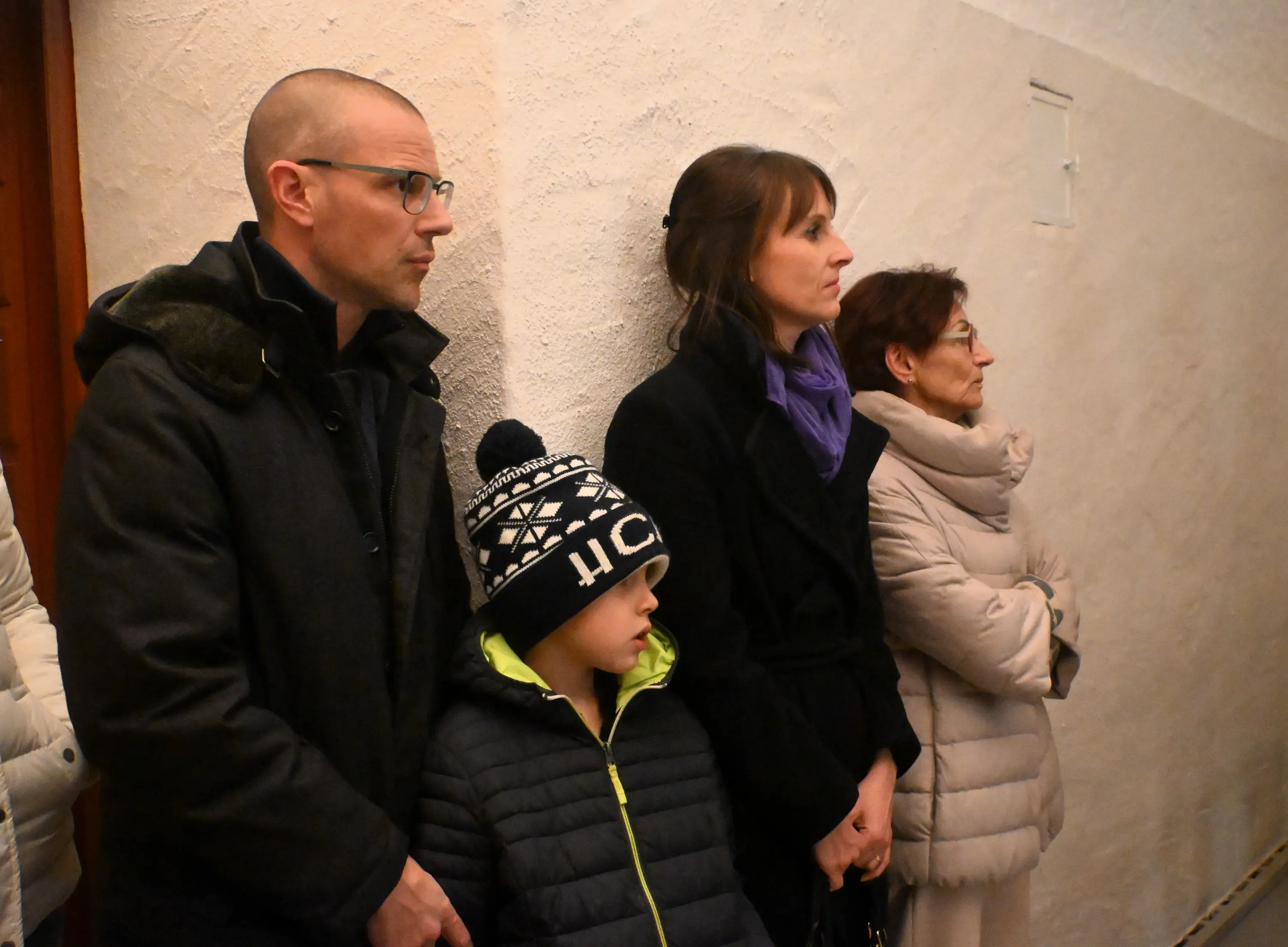 Un gruppo di persone in piedi, in un ambiente interno. Un uomo con occhiali e giacca scura, una donna con sciarpa viola, un bambino con cappello a righe