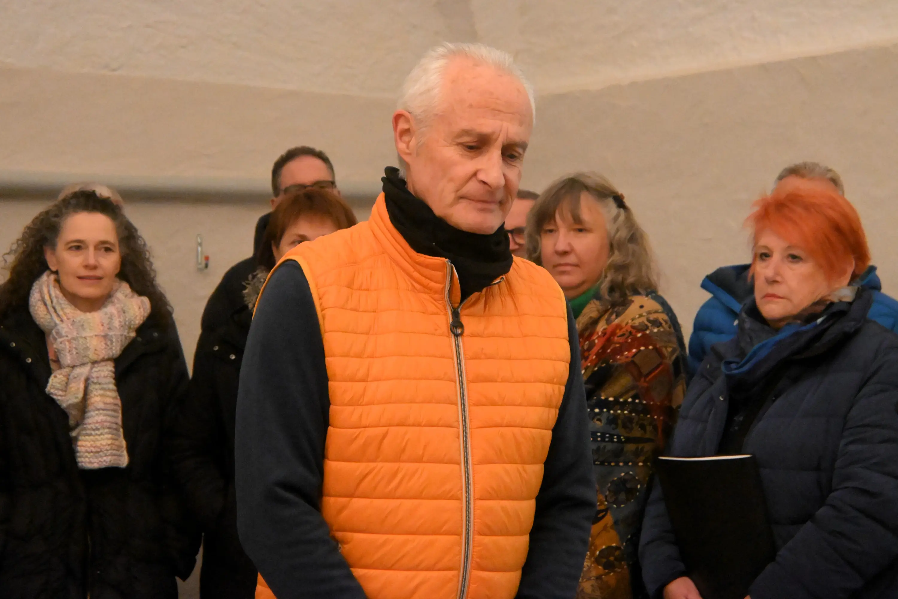 Un uomo in giacca arancione è in primo piano, visibilmente serio. Dietro di lui, un gruppo di persone di varie età e stili, alcune con sciarpe