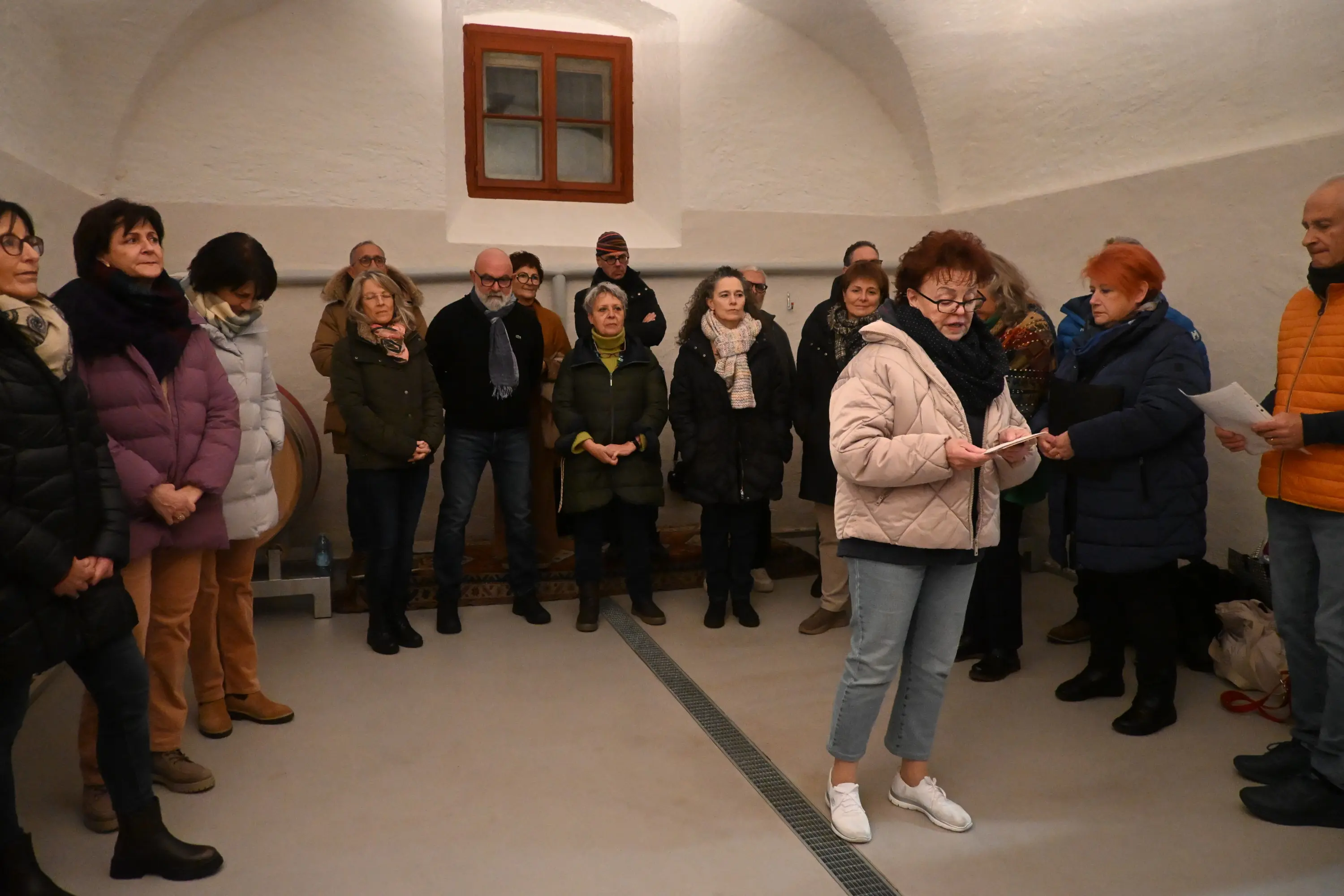 Un gruppo di persone si riunisce in un ambiente interno, ascoltando un discorso. Alcuni partecipanti sono in piedi in cerchio, mentre una donna con un giubbotto