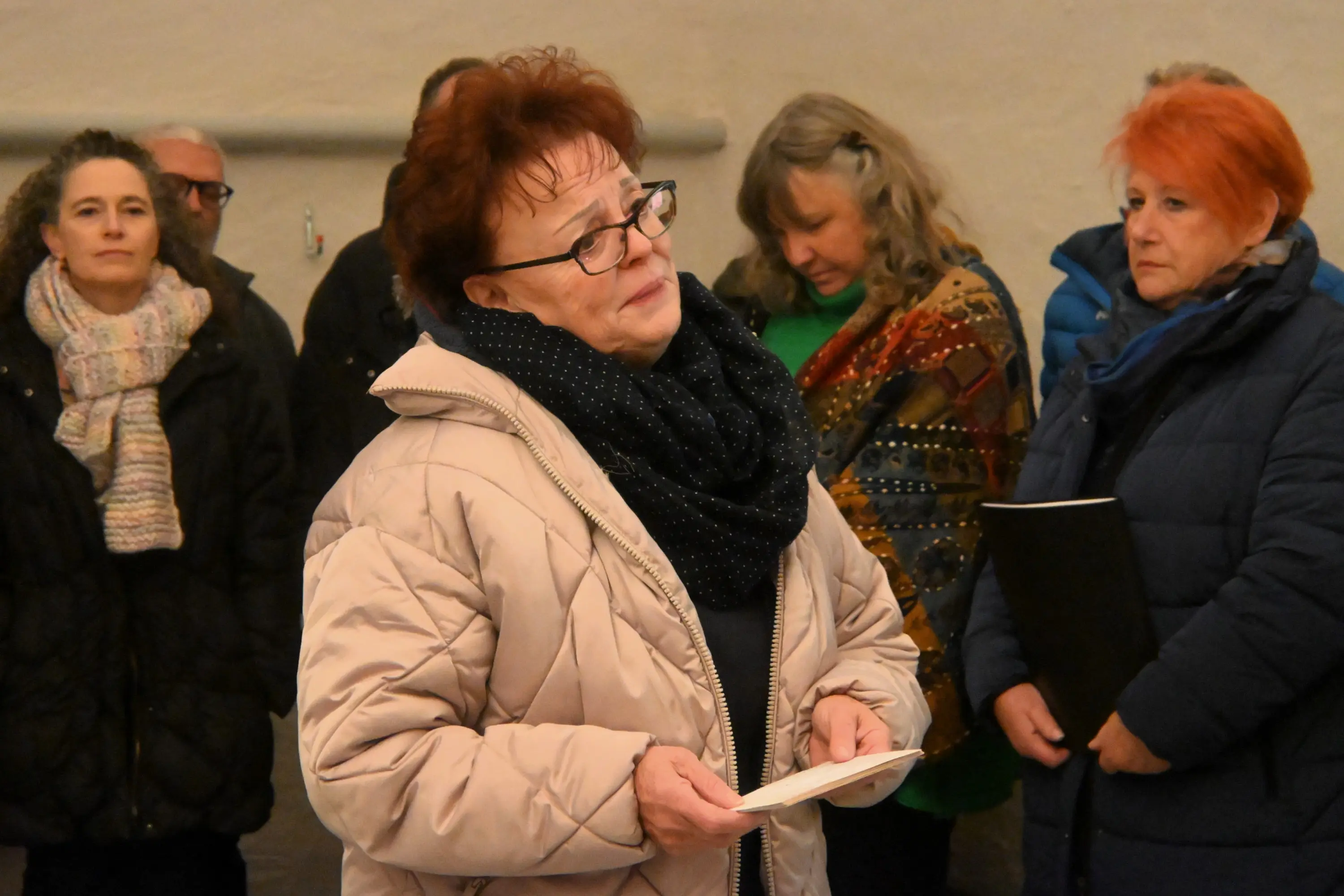 Una donna con capelli rossi e occhiali, indossando un giubbotto beige, legge un testo mentre un gruppo di persone la osserva attentamente in un ambiente interno. Alc