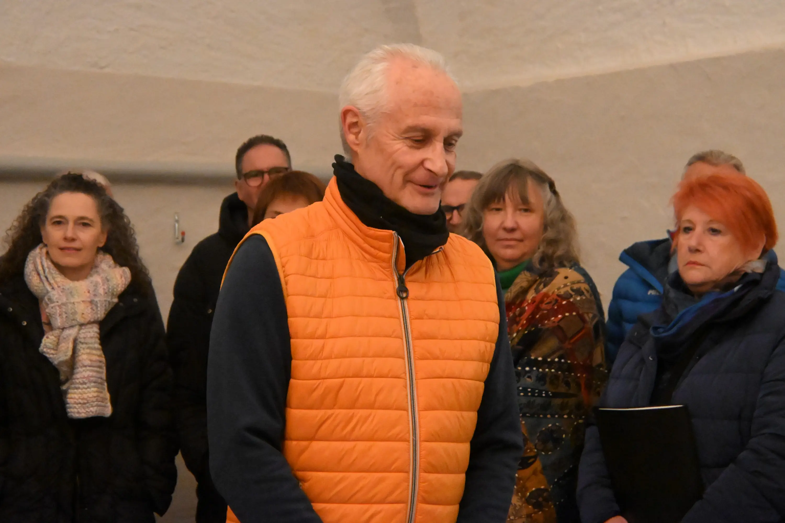 Un gruppo di persone si trova in una stanza, con un uomo al centro che indossa un gilet arancione e sorride. Le donne e gli uomini sullo sfondo sono in ab