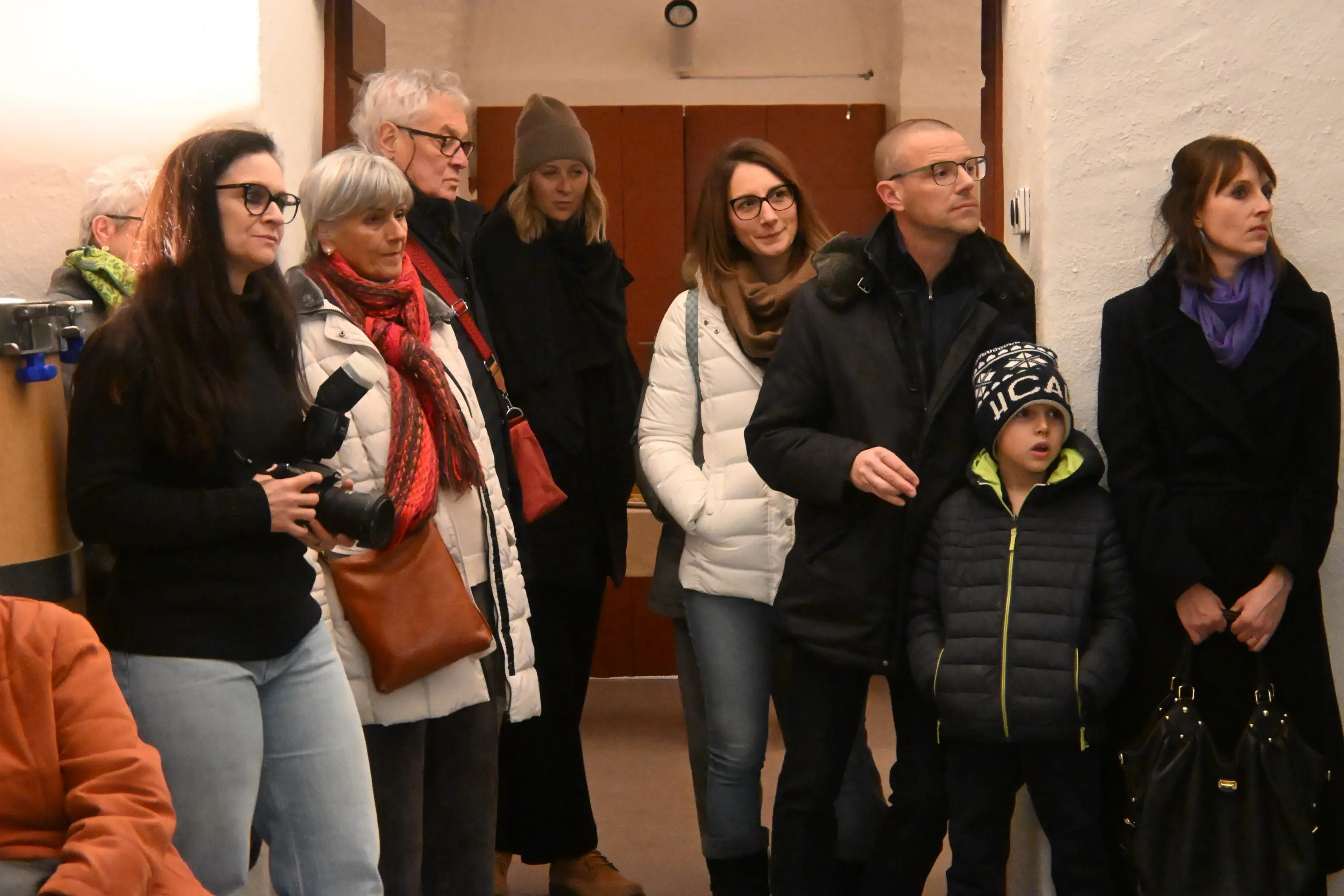 Un gruppo di persone in un ambiente interno, alcune con giacche bianche e sciarpe. Alcuni adulti stanno osservando, mentre un bambino è al centro del gruppo. L