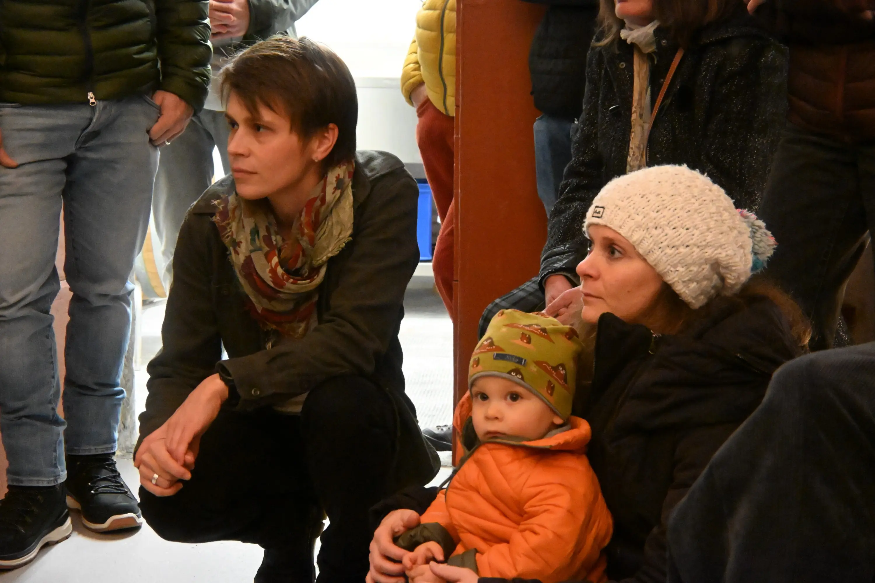 Gruppo di persone sedute, con due adulti e un bambino. Un adulto indossa una sciarpa e sembra ascoltare attentamente, mentre l'altro adulto tiene in braccio il