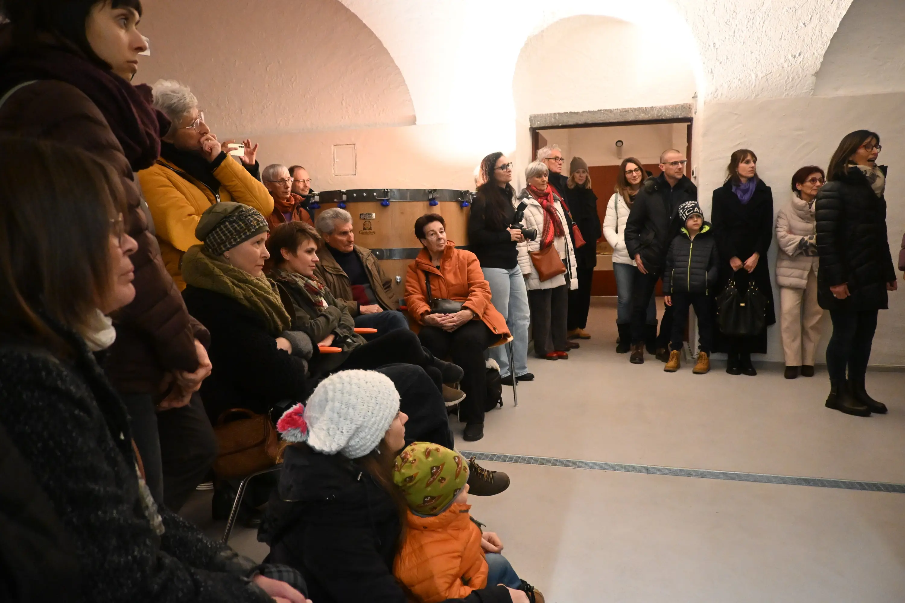 Una folla di persone sedute e in piedi in una sala con pareti chiare e archi, alcune persone con cappelli e sciarpe, attentamente in ascolto.