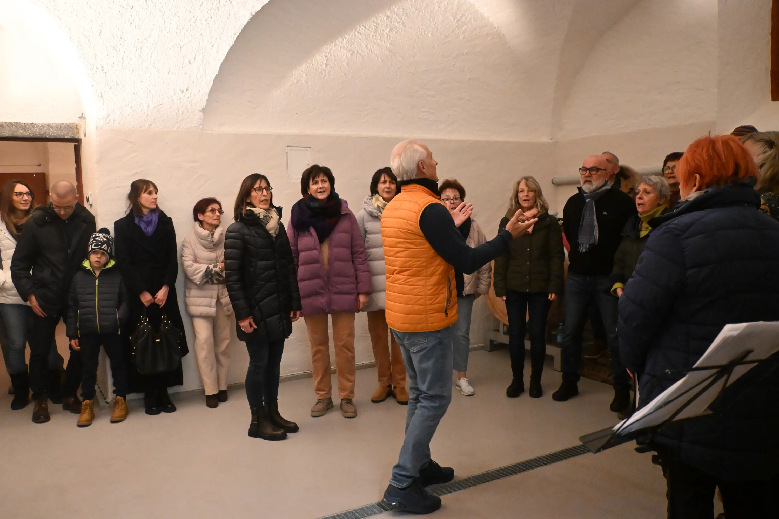 Un gruppo di persone, vestite in giacche invernali, canta insieme in una sala. Un uomo con un gilet arancione si trova di fronte al gruppo, guidando l