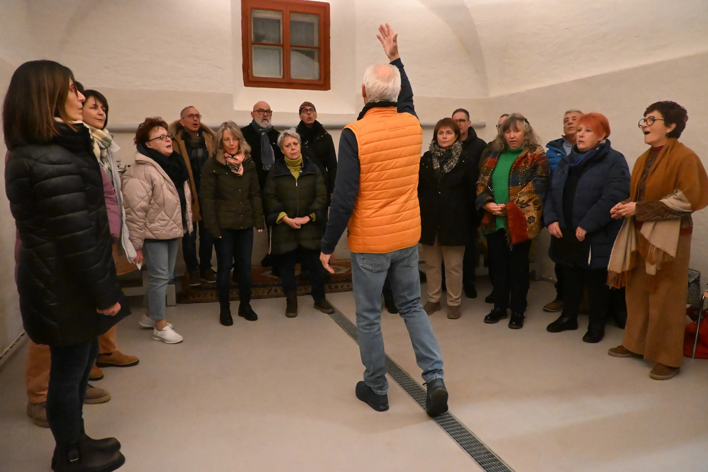 Un gruppo di persone si riunisce in una sala, mentre un uomo in piedi con una giacca arancione alza una mano per dirigere. I partecipanti, di diverse età