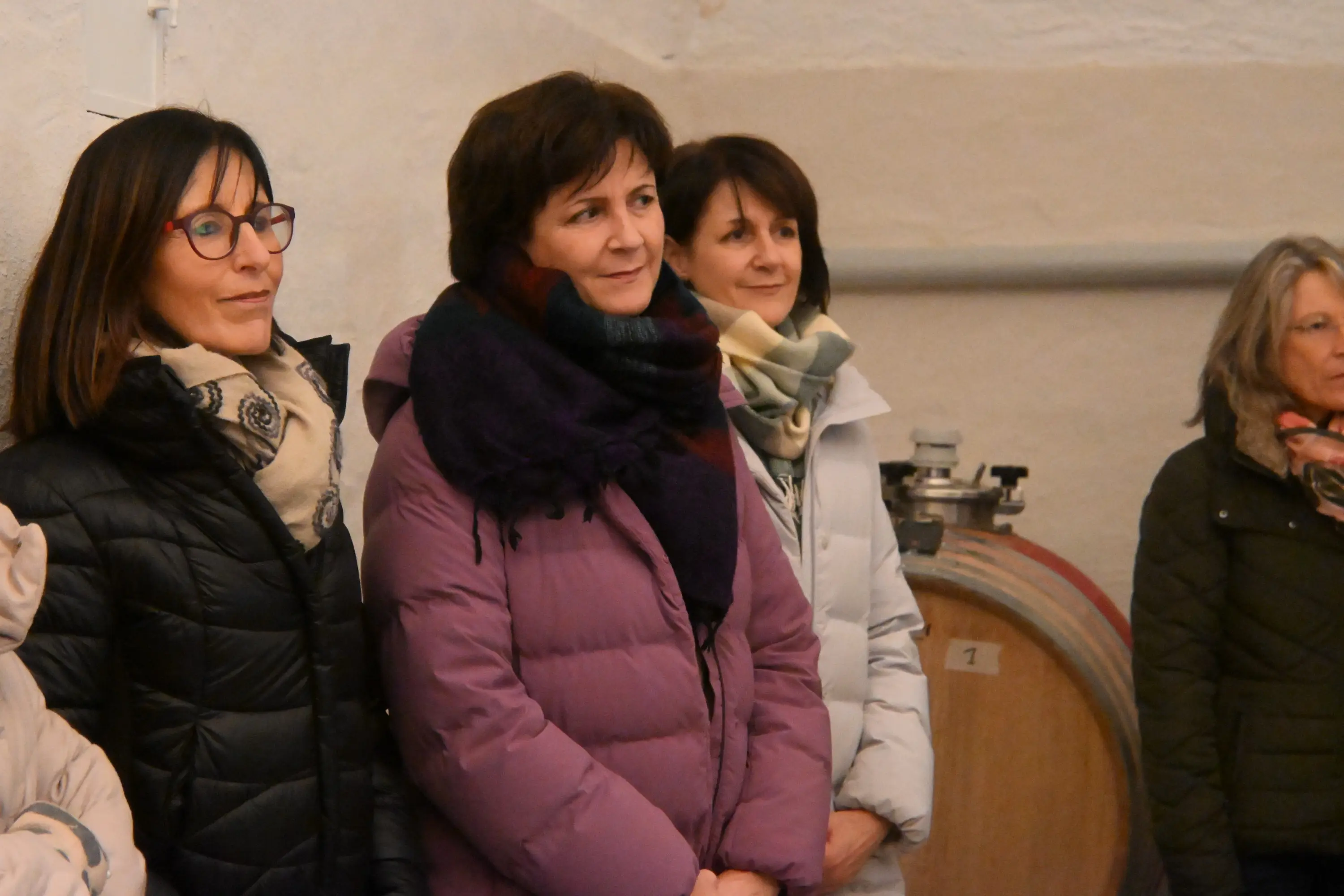 Un gruppo di donne, vestite con giacconi e sciarpe, ascolta attentamente in una cantina. Sullo sfondo si vedono botti di legno.