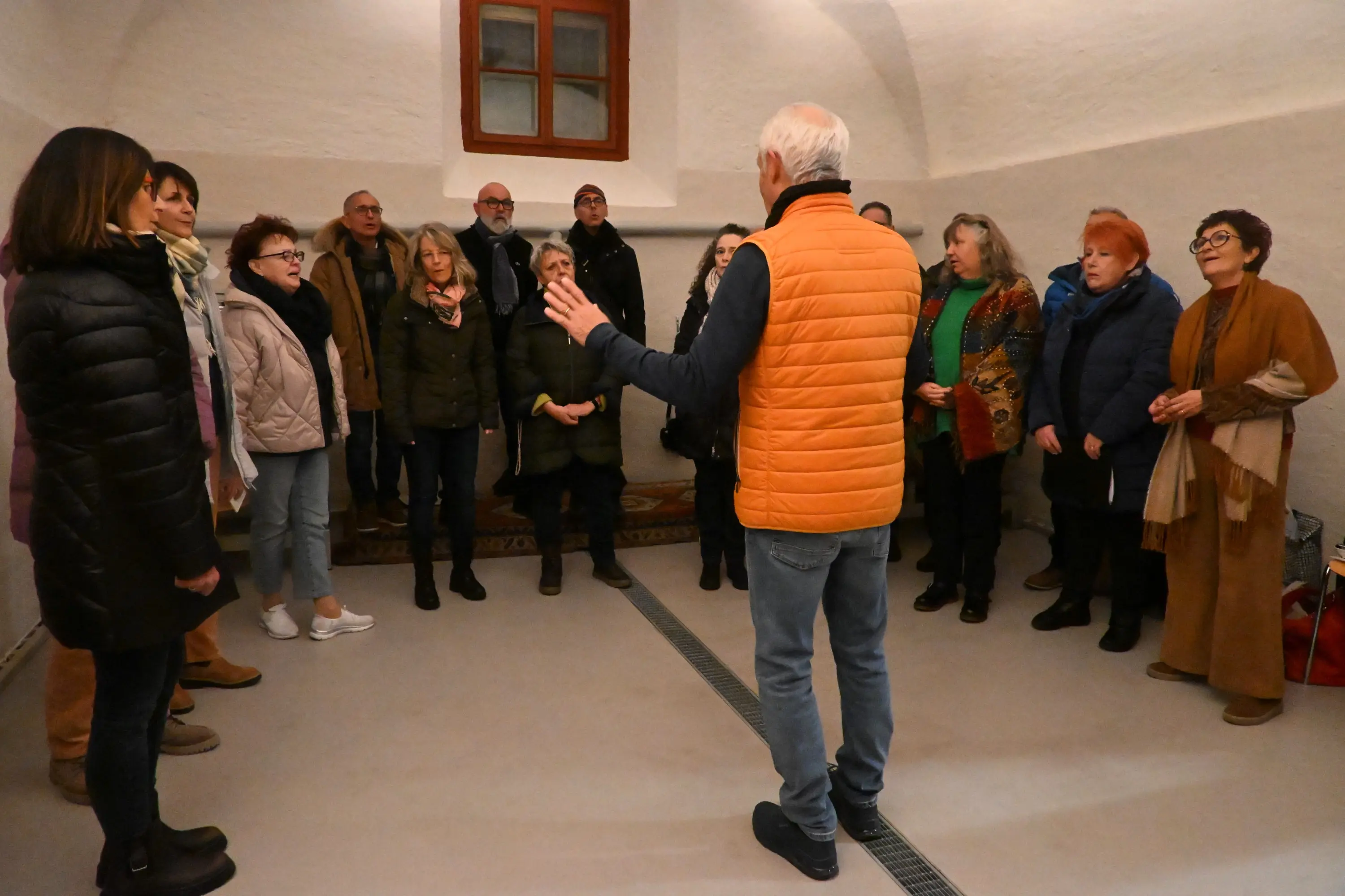 Un gruppo di persone di varie età si trova in una stanza con pareti bianche. Un uomo con un gilet arancione guida l'assemblea, mentre i membri del coro