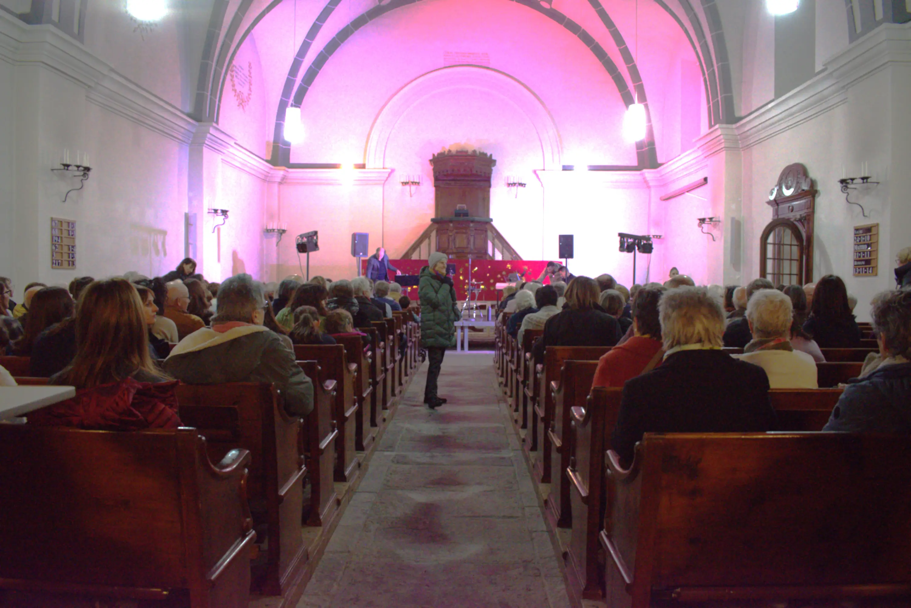 Interno di una chiesa affollata con gente seduta e in piedi, illuminata da luci rosa. Un uomo in piedi si dirige verso l'altare.