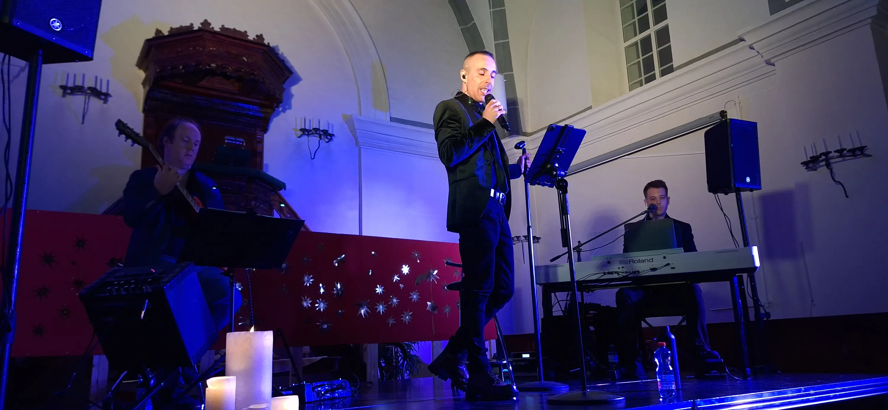 Un uomo canta al microfono su un palco, mentre due musicisti lo accompagnano con un pianoforte e chitarra. L'illuminazione è blu e ci sono candele