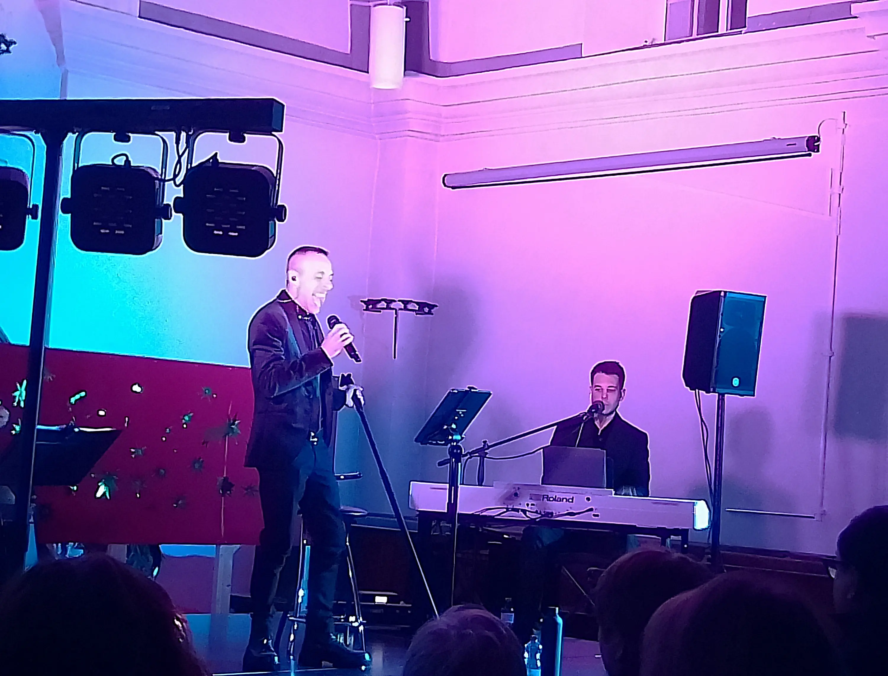 Un cantante si esibisce sul palco, con un microfono in mano, mentre un musicista lo accompagna al pianoforte. L'illuminazione è colorata, con tonalità