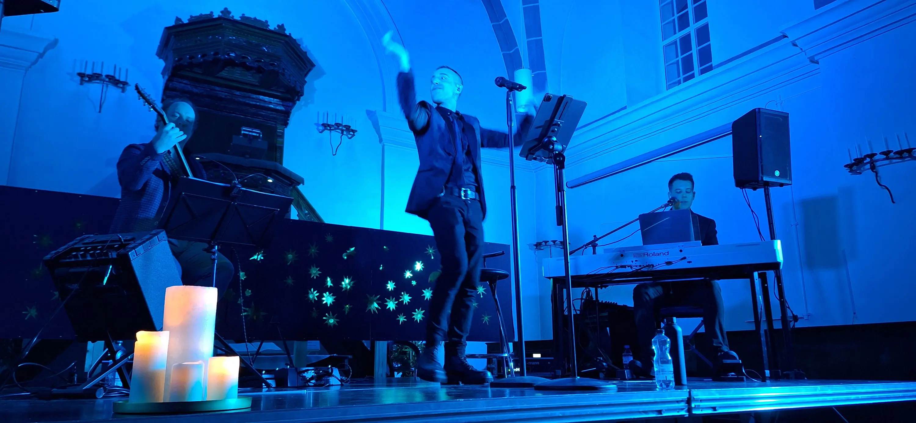 Un musicista in abito elegante si esibisce su un palco illuminato da luci blu, mentre due musicisti lo accompagnano, uno alla chitarra e l'altro al pian