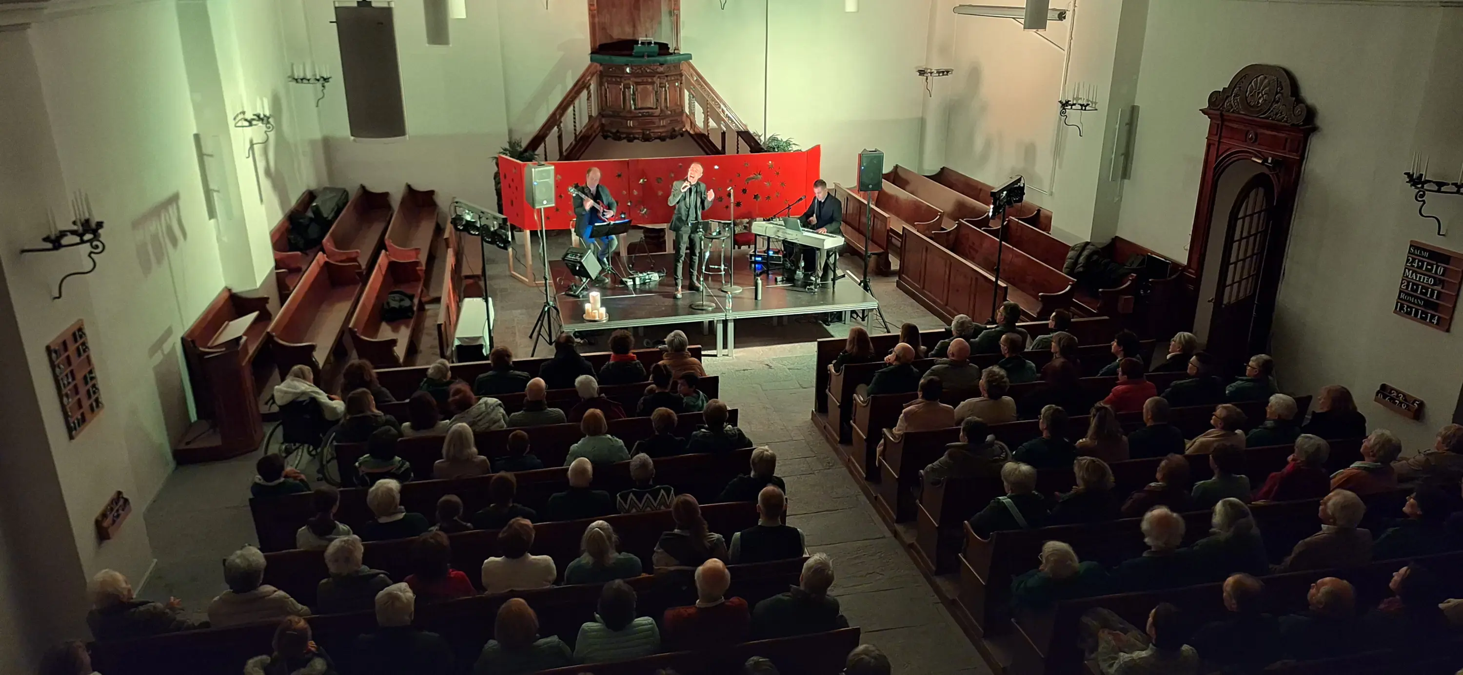 Un concerto si svolge in una chiesa con un pubblico numeroso. Sul palco, quattro musicisti suonano strumenti mentre un grande fondale rosso è decorato con motivi floreali