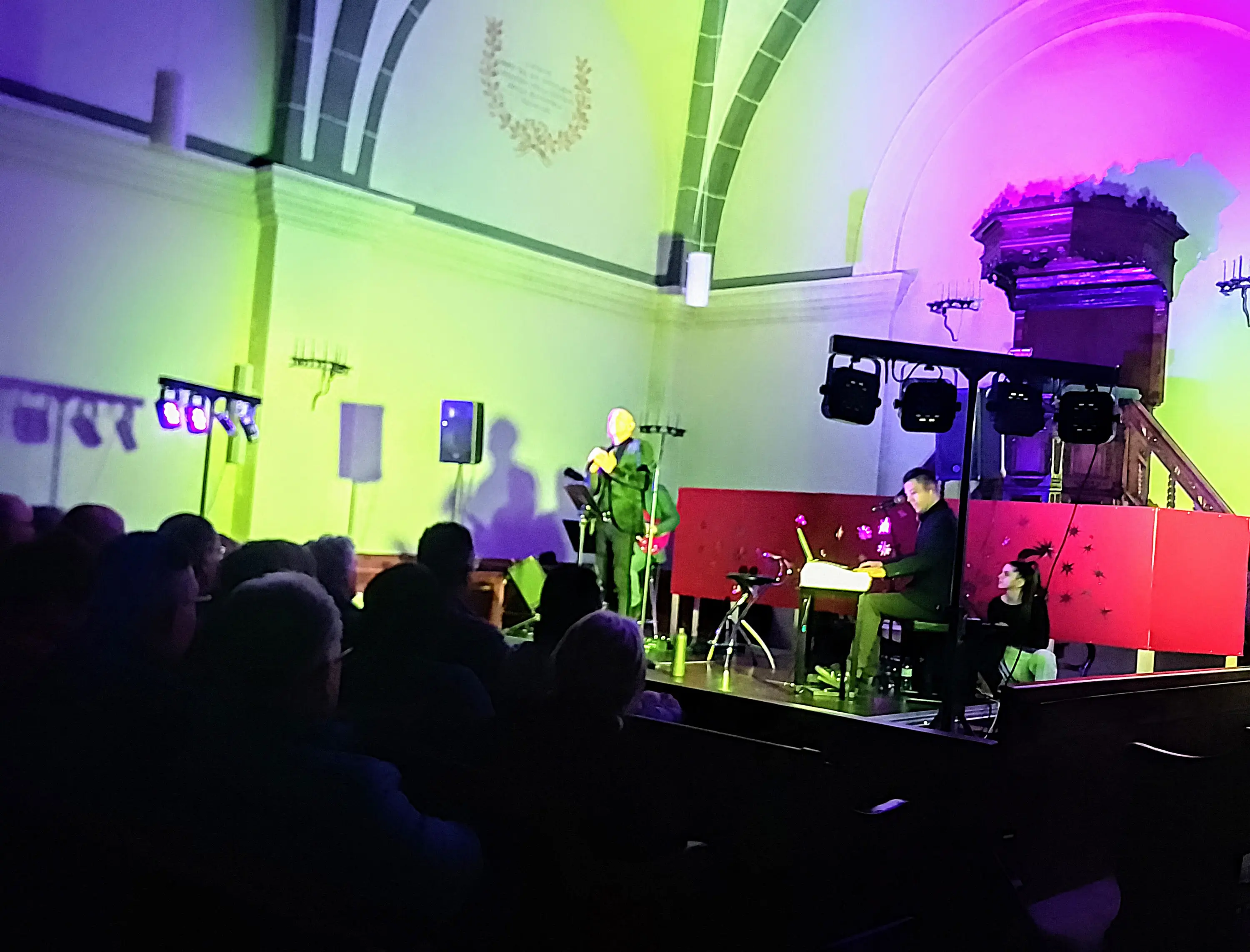 Un performer su un palco illuminato da luci colorate, mentre un pianista suona di spalle. Il pubblico, seduto e in attesa, osserva attentamente. Sullo