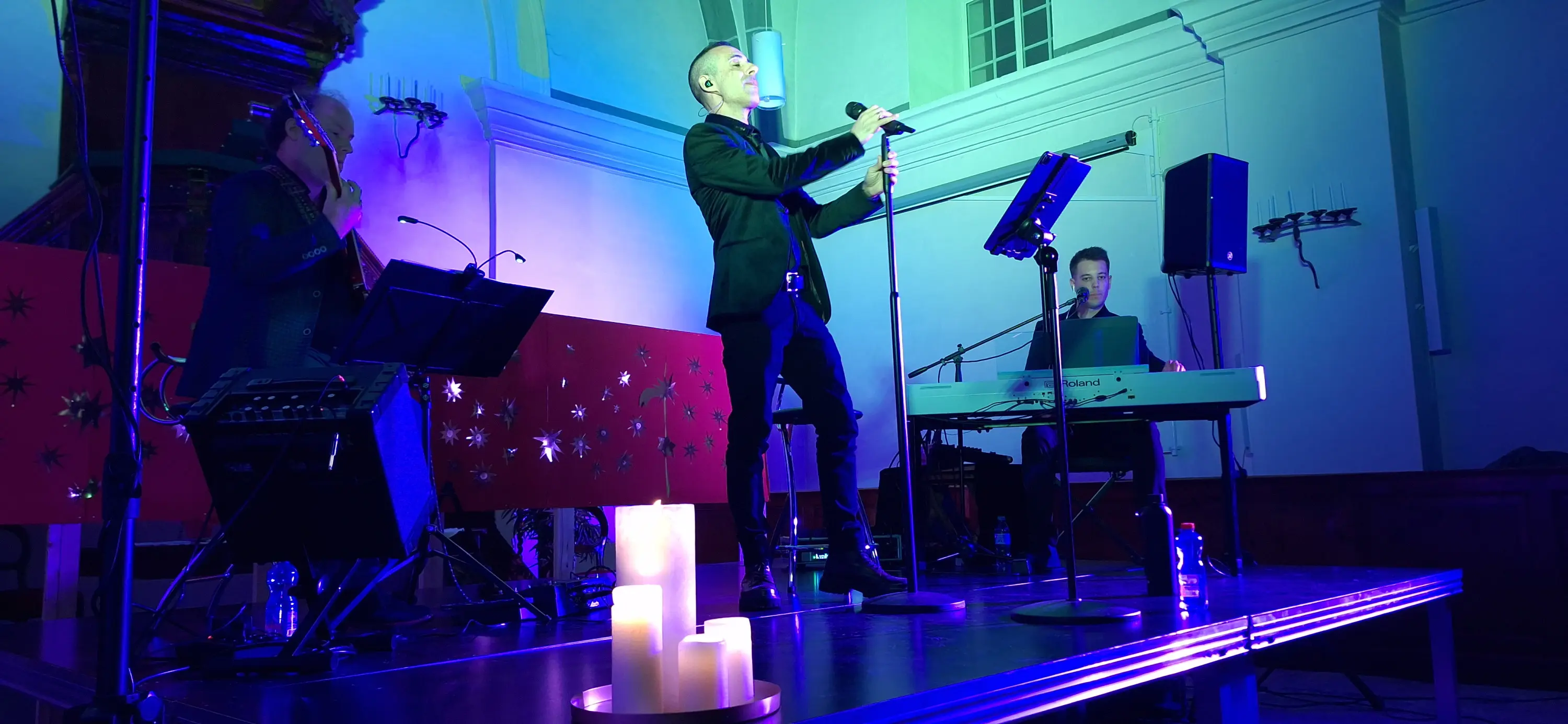 Un artista in abito elegante canta sul palco, con un microfono in mano. Sullo sfondo, due musicisti suonano strumenti. L'atmosfera è illuminata da cande
