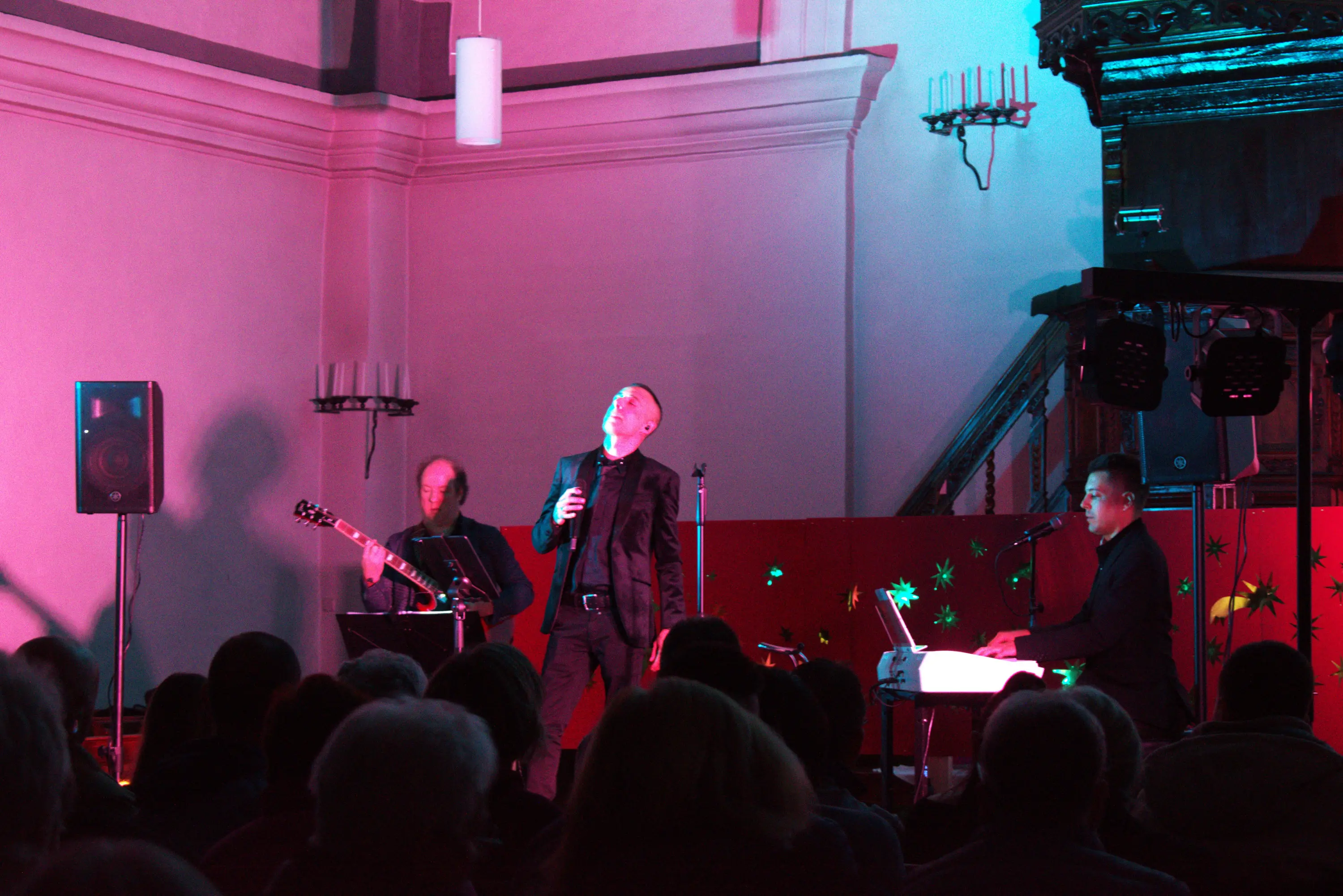 Un uomo canta sul palco, mentre un chitarrista e un pianista lo accompagnano. La stanza è illuminata con luci colorate e il pubblico è visibile in prima fila.