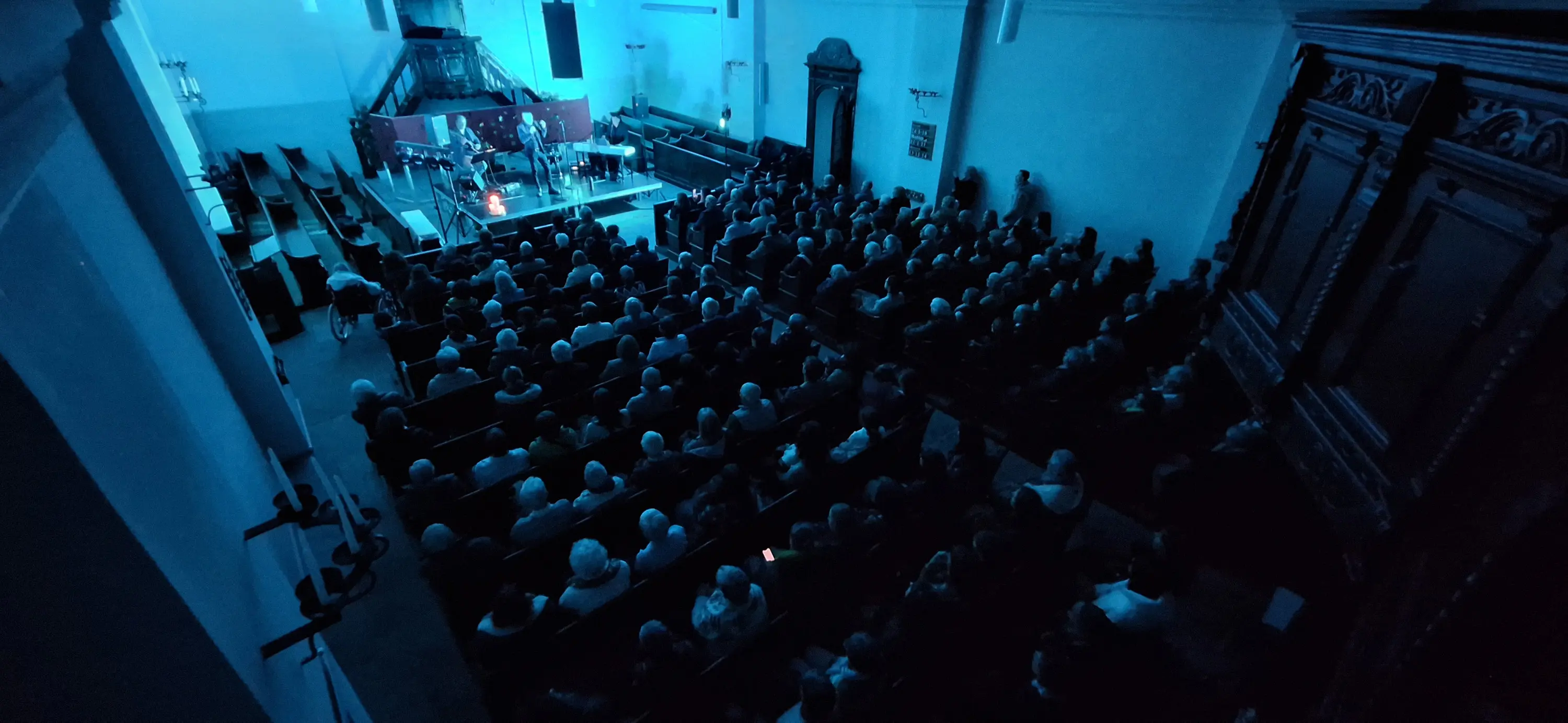 Una sala gremita di persone sedute, illuminate da una luce blu durante un evento, con musicisti sul palco visibili in lontananza. Le panche di legno e l'arch
