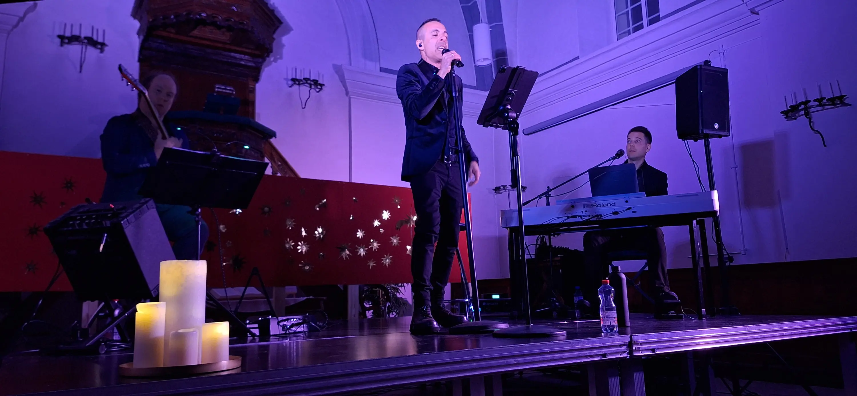 Un uomo canta sul palco, indossando un abito elegante. Dietro di lui, un musicista suona la chitarra e un altro è al piano. Sulla sinistra,