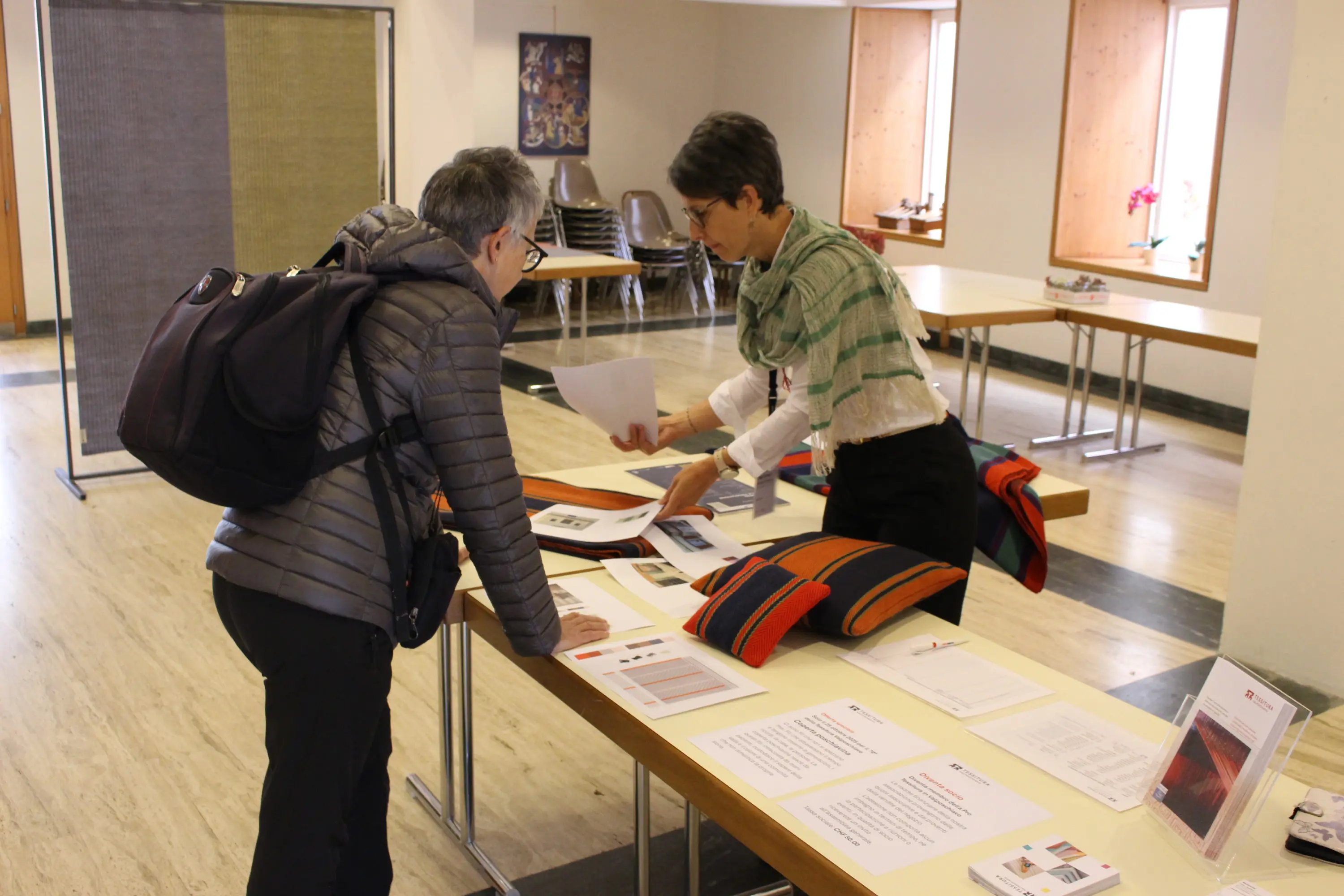 Due persone in un ambiente espositivo. Una donna, con occhiali e un copricapo scuro, esamina documenti su un tavolo. L'altra donna, con un foulard e capelli corti, le mostra materiali promozionali. Sul tavolo ci sono vari opuscoli