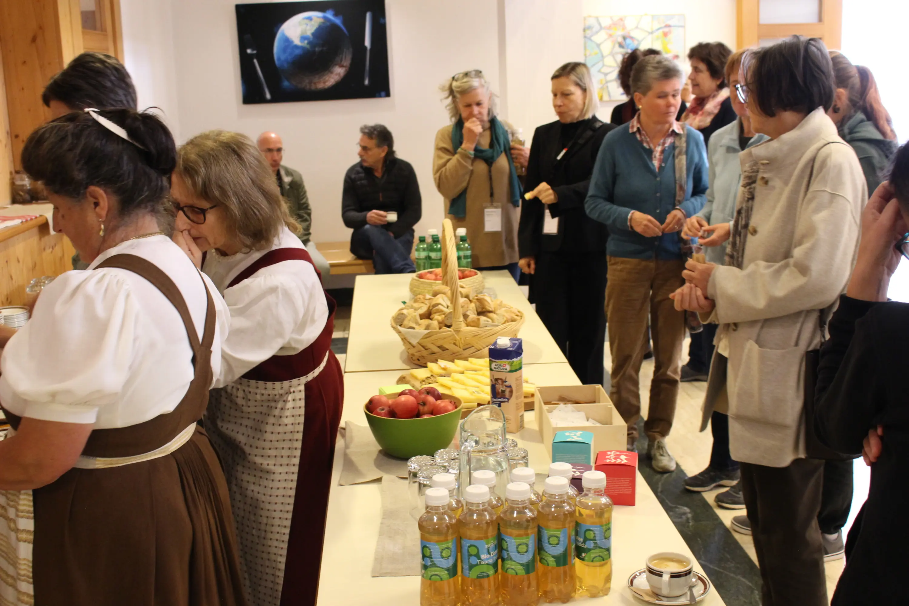 Un gruppo di persone si riunisce in una sala, alcuni chiacchierano mentre altri si servono da un tavolo imbandito con cibo e bevande. Sullo sfondo, è visibile un monitor che mostra un'immagine della Terra.