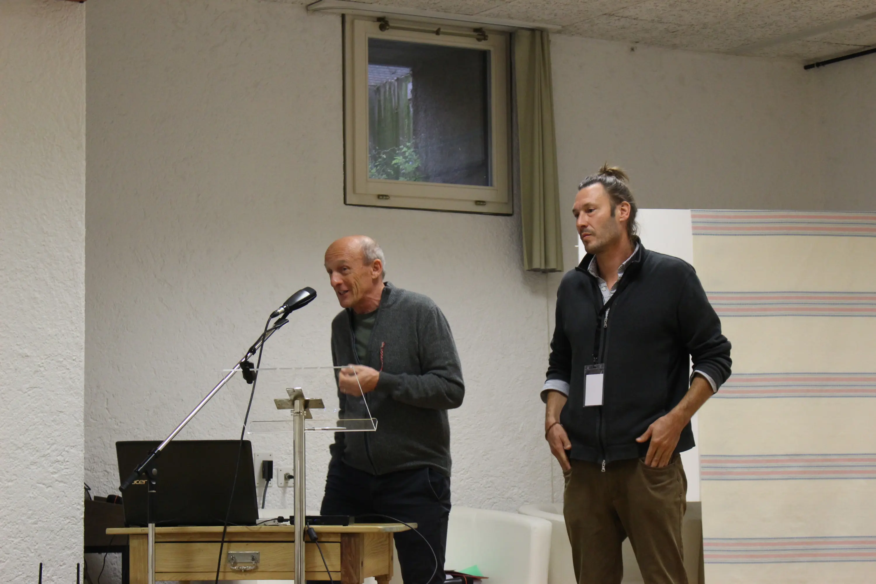 Due uomini parlano a un pubblico in un ambiente interno. Uno sta al microfono mentre l'altro lo ascolta attentamente. Sul tavolo ci sono un computer portatile e un leggio.