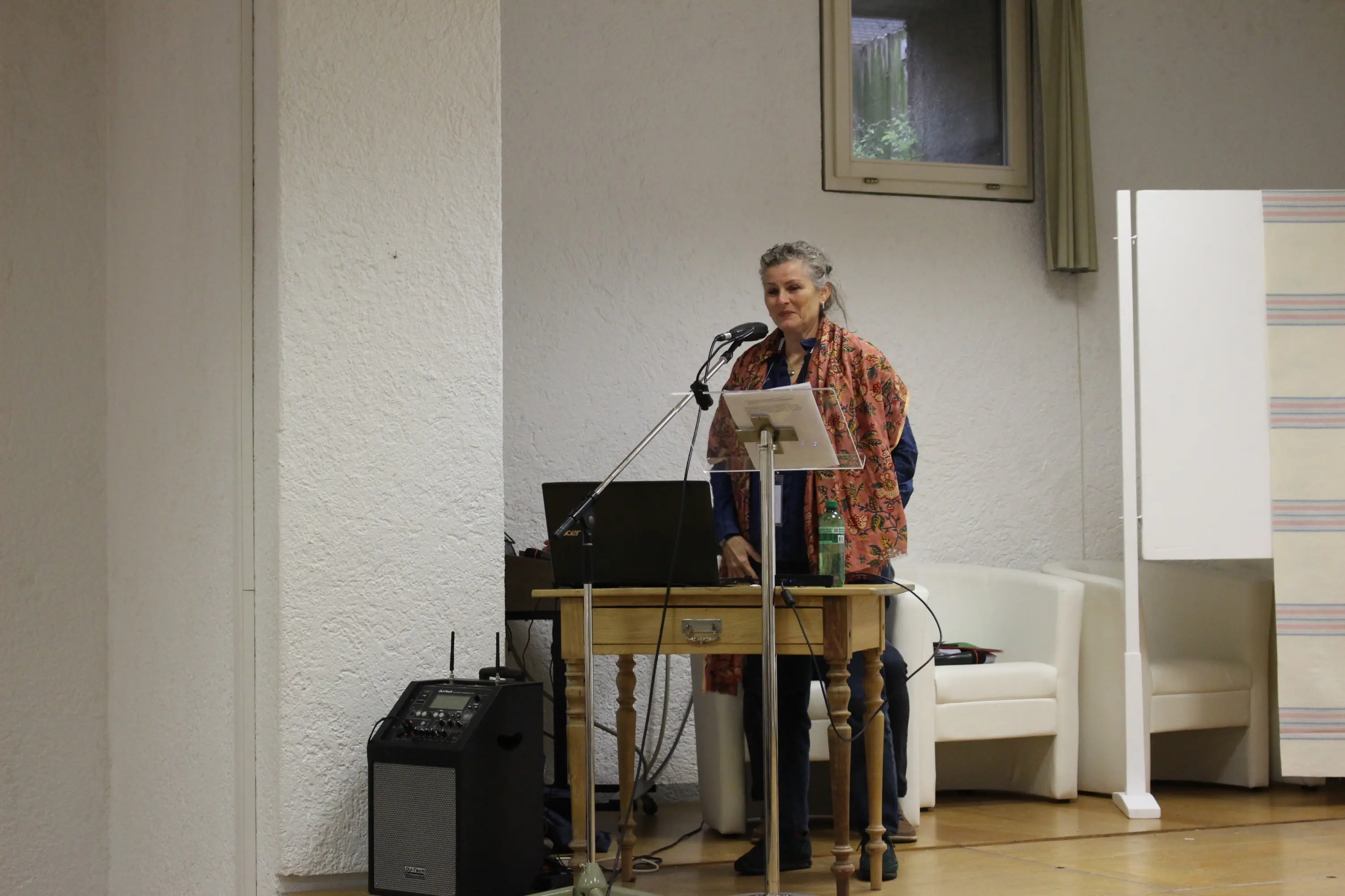 Una donna con capelli corti e grigi parla a un microfono, con un tavolo e un computer portatile davanti a lei. Indossa una sciarpa colorata e sembra essere a un evento o a una conferenza in una sala bianca.