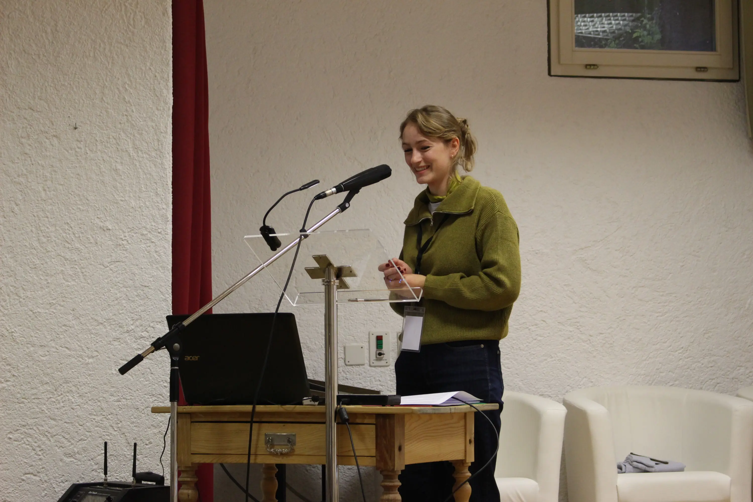 Una giovane donna con capelli biondi raccolti sorride mentre parla a un podio trasparente. Indossa un maglione verde e jeans, con un computer portatile sul tavolo di fronte a lei. Sullo sfondo si vedono pareti bianche e una t