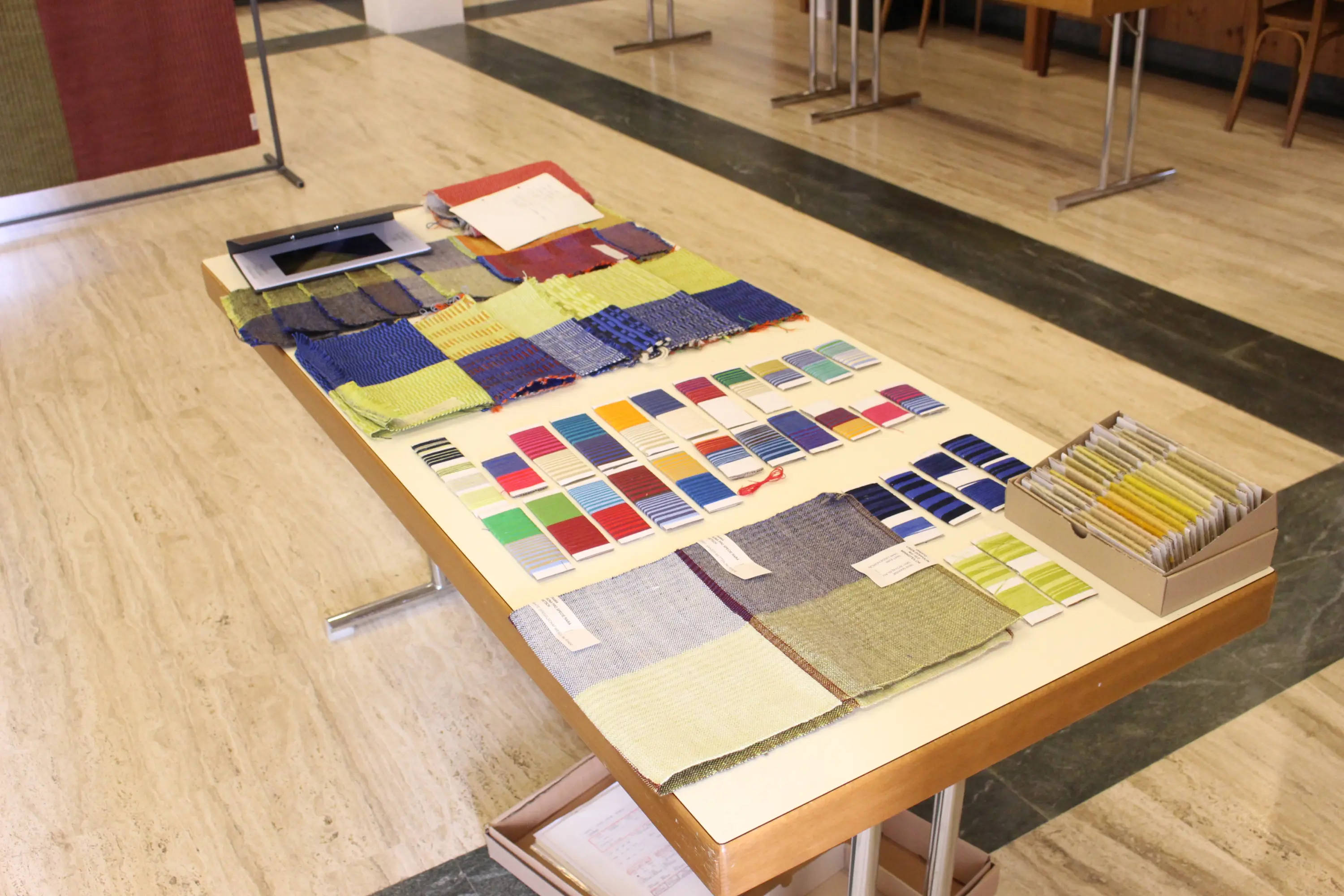 Tavolo con diversi campioni di tessuti e schede di colori, organizzati in una presentazione di materiali variopinti, su un pavimento di marmo in un ambiente luminoso.