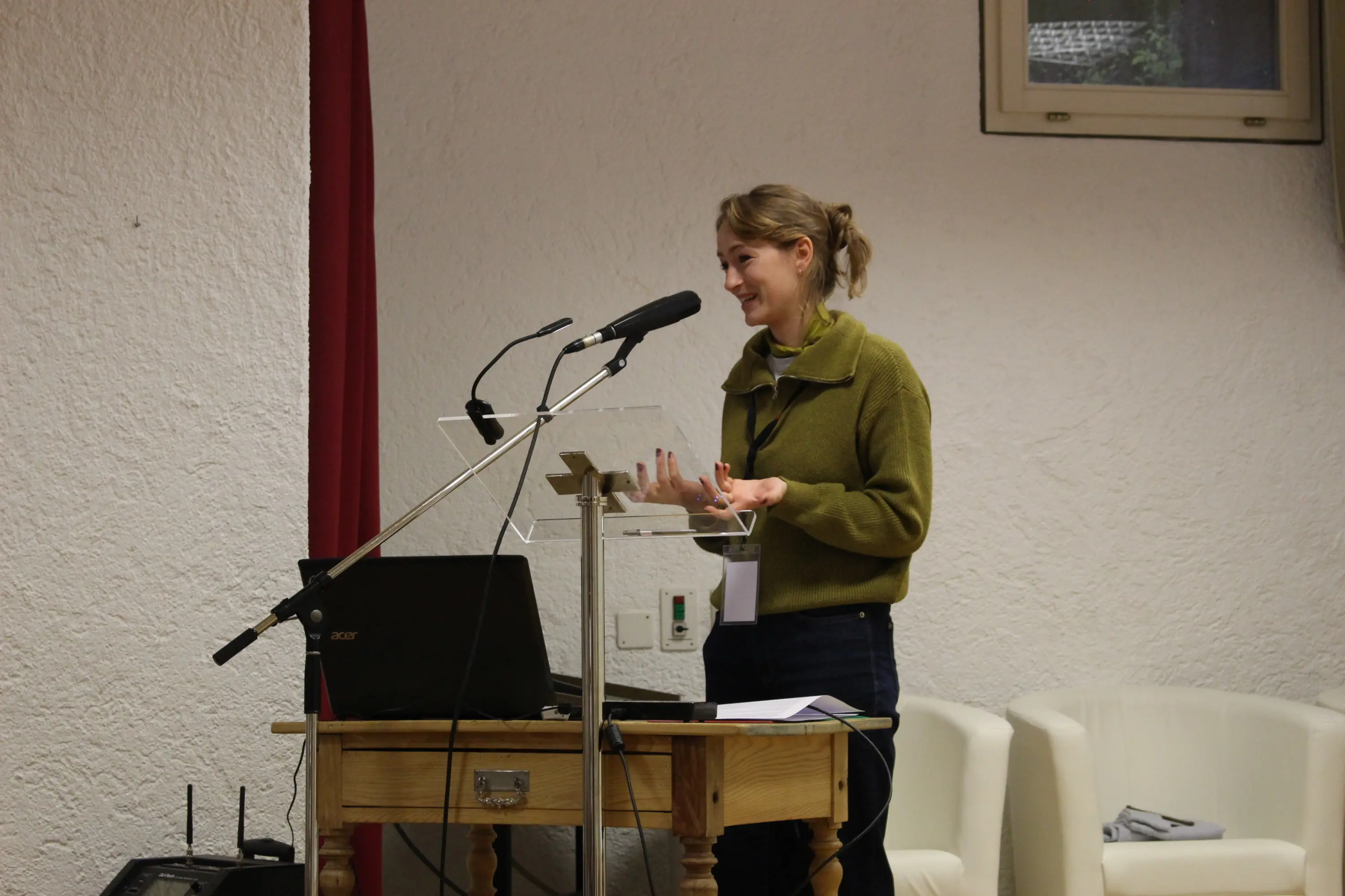 Una donna sorride mentre parla a un podio con microfoni durante una presentazione. Indossa un maglione verde e jeans. Sul tavolo ci sono un computer portatile e dei documenti. Sullo sfondo, una parete bianca e alcuni arredi.