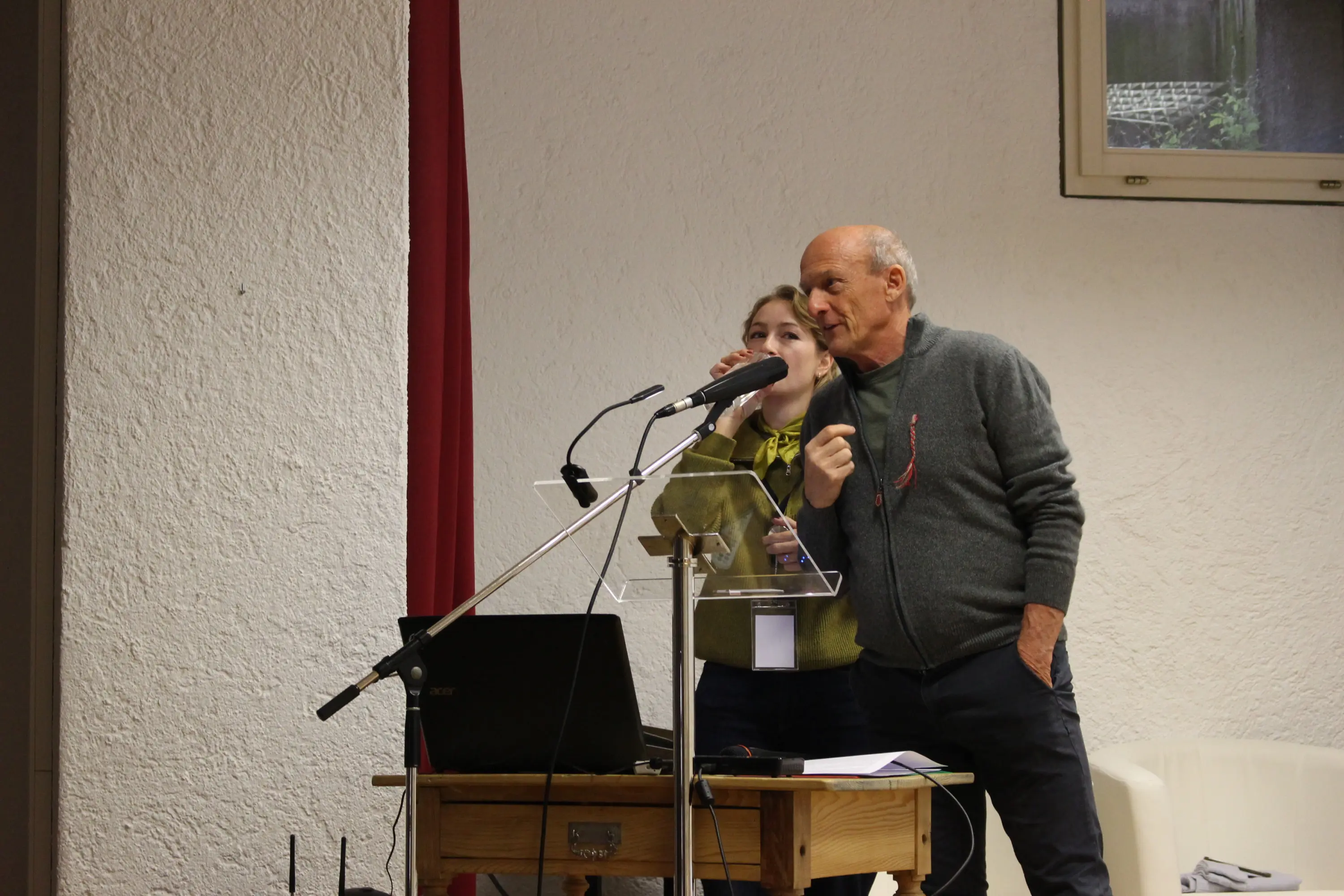 Due persone, un uomo e una donna, sono davanti a un microfono e un computer portatile. L'uomo parla con passione, mentre la donna lo assiste e tiene un microfono. Sullo sfondo, una parete bianca e un'illuminazione soft.